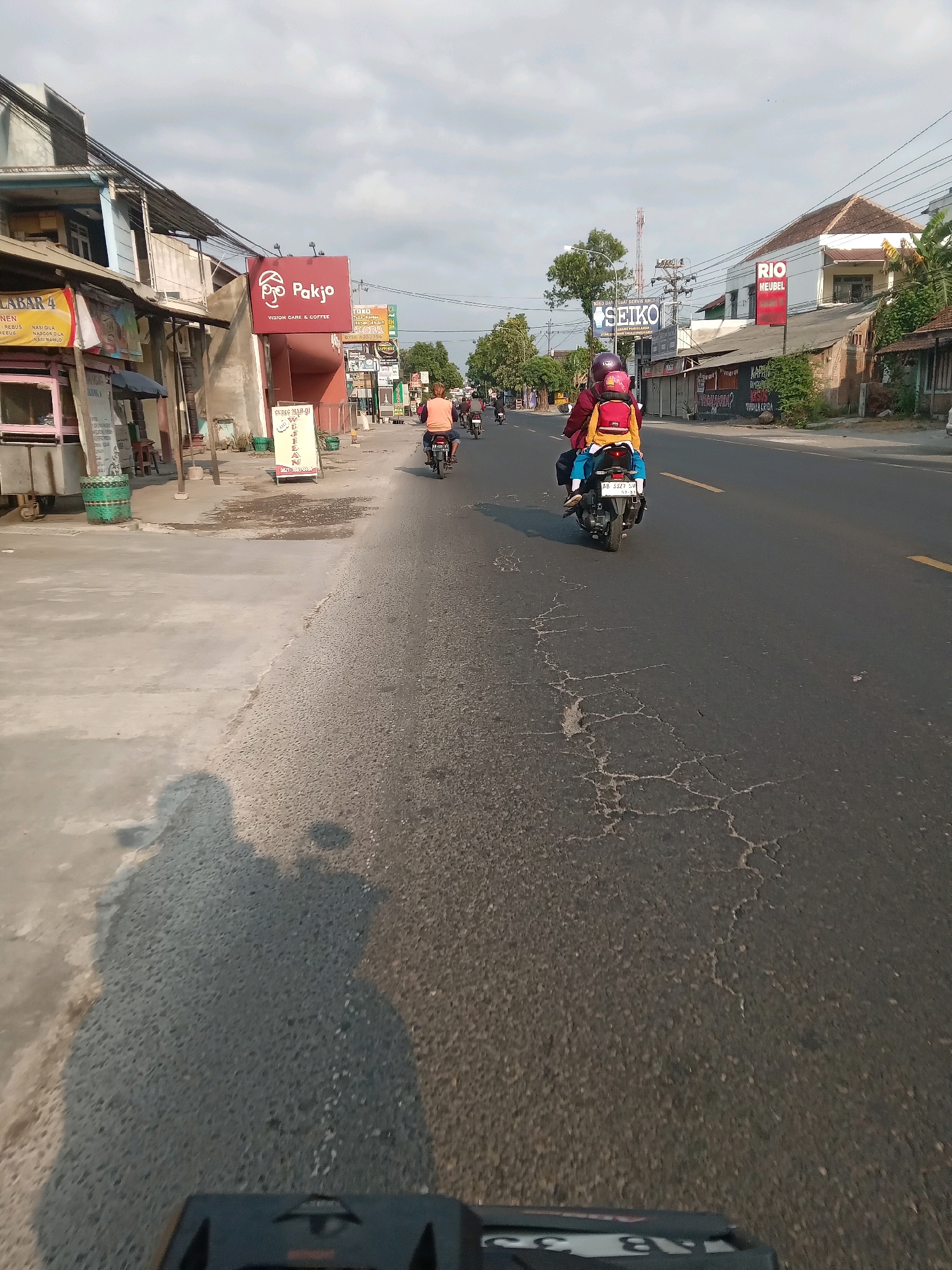 This is a street-level, forward-facing shot of a road in Sitimulyo, Indonesia, taken from the perspective of a moving vehicle, likely a motorcycle, given the shadow in the foreground. The road is asphalt with some cracks and a yellow line marking the center. On the left side of the road, there are several buildings with storefronts, including one with a prominent red sign displaying "Pakjo VISION CARE & COFFEE". There are also other signs for businesses like "Seiko" and "Rio Meubel". Several motorcycles are visible on the road, some carrying passengers. One motorcycle in the mid-ground has two people on it, a driver and a passenger wearing a yellow backpack. The sky is overcast with some visible clouds. The overall scene depicts a typical urban or semi-urban street environment in Indonesia.