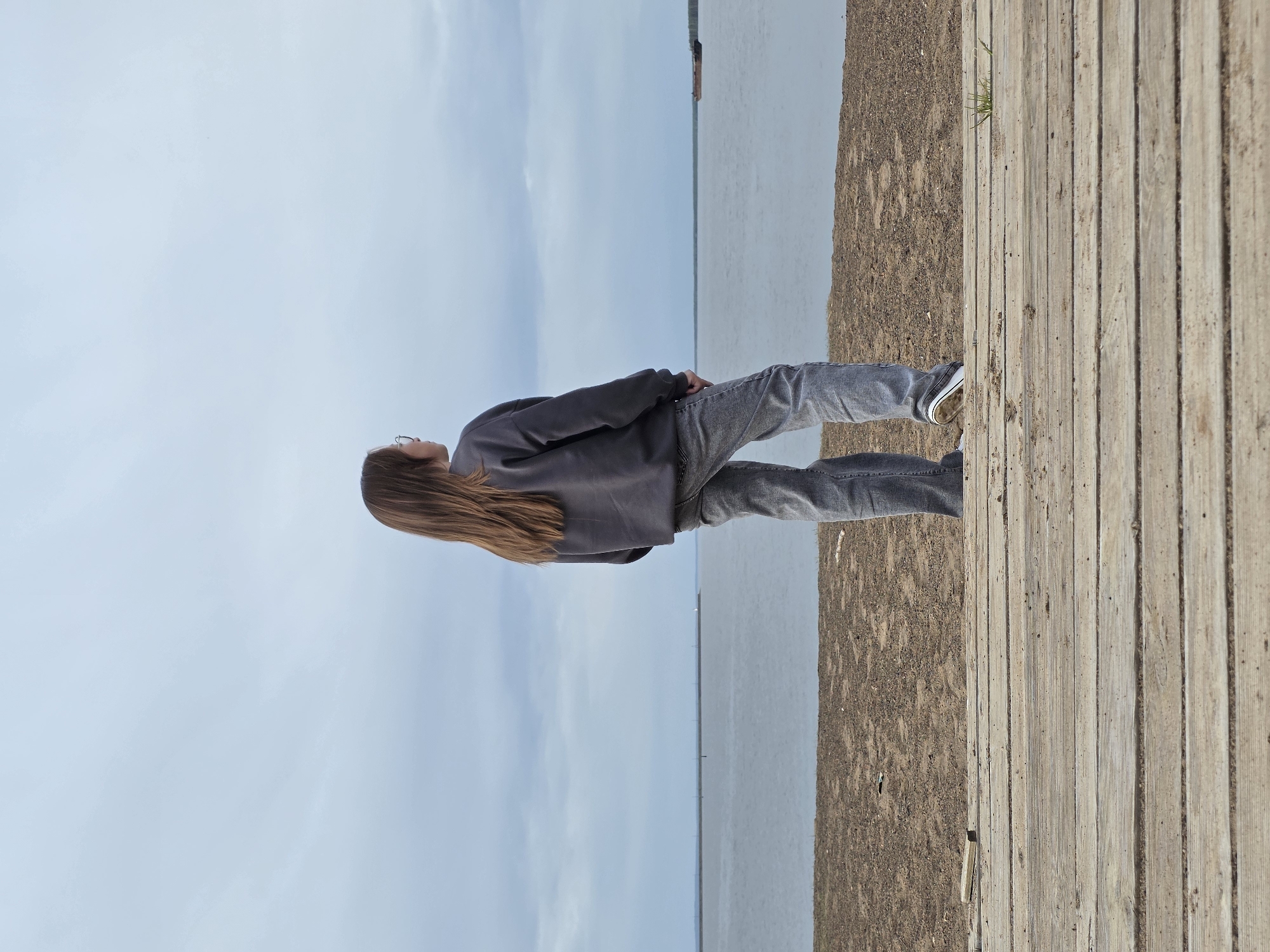 A young person with long brown hair, wearing a gray sweatshirt and gray jeans, stands looking up in a vertical orientation on a boardwalk next to a sandy area and a body of water under a cloudy sky. This location is in Chaikovsky, Russia.