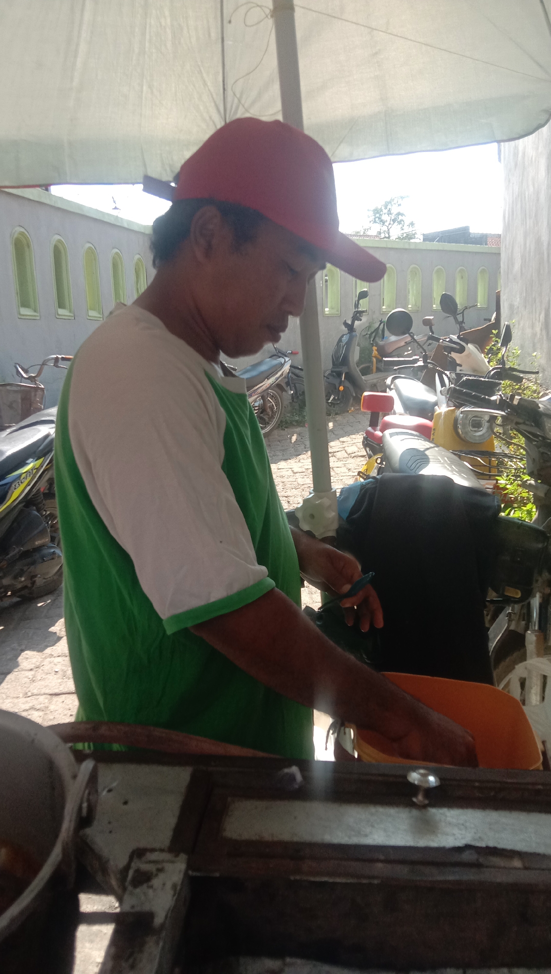 A man wearing a red baseball cap and a green and white t-shirt is standing outdoors in the city of Karang Anyar, Indonesia. He is leaning over a cart, preparing food. Behind him, several motorcycles are parked, and a light-colored building with arched windows is visible. The man is holding a small blue utensil in his right hand. The scene appears to be a street-side food stall, with the man actively engaged in his work.