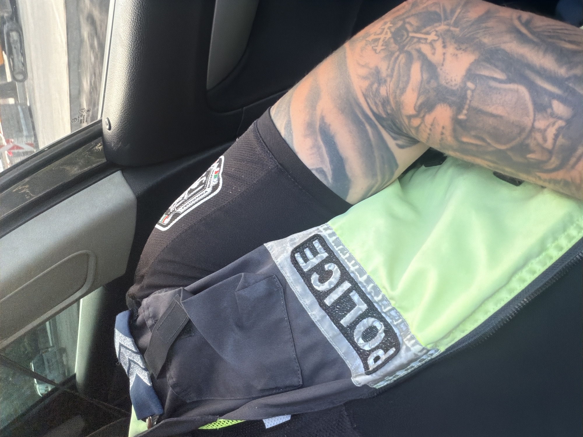An interior shot from within a Volkswagen passenger vehicle depicts the right arm and upper torso of an individual. The individual wears a dark-colored, short-sleeved police uniform shirt featuring a patch on the right sleeve with the text "РЕПУБЛИКА БЪЛГАРИЯ" (Republic of Bulgaria) and "ПОЛИЦИЯ" (Police). A high-visibility vest or garment, partially visible over the uniform shirt, displays the word "POLICE" in reflective lettering on a dark background above a neon green lower section. The individual's right arm is heavily tattooed, primarily with a large, roaring lion's face and a cross-like symbol near the lion's head, covering the forearm and extending onto the upper arm. Through the vehicle's window, an outdoor urban scene during daytime is visible, showing a road or construction site with orange and white traffic barriers and a tracked excavator in the background. The location is an urban area within Shumen, Bulgaria, characterized by active road construction.