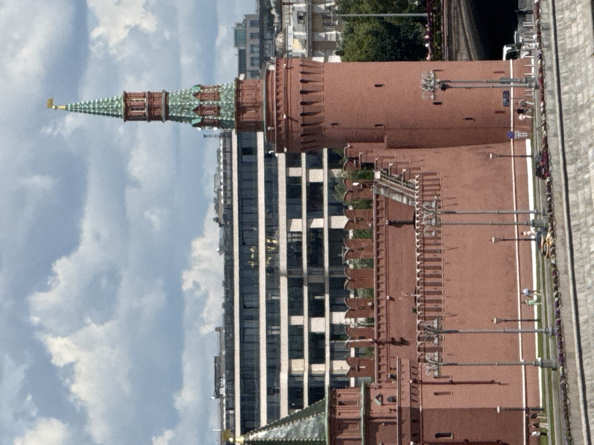 This is a vertically oriented, medium-close shot of a red brick tower with a green spire, characteristic of Russian architecture, set against a backdrop of a cloudy sky. The tower appears to be part of a larger fortress or complex, with battlements visible along its upper edge. A modern, multi-story building with a striped facade of white and dark windows stands to the left of the tower, partially obscuring it. Below the tower, more of the red brick structure is visible, featuring arched architectural details. In the foreground, a walkway or street is visible, with traffic lights and street lamps lining its edge. A few figures, too small to discern details, can be seen on this walkway, suggesting it's a public area. The overall scene depicts an urban environment with historical and modern elements, likely taken during daylight. The weather appears partly cloudy. The image's orientation suggests it was taken with the camera held vertically. The location cues and architectural style strongly indicate Moscow, Russia. No visible text is present in the image.
