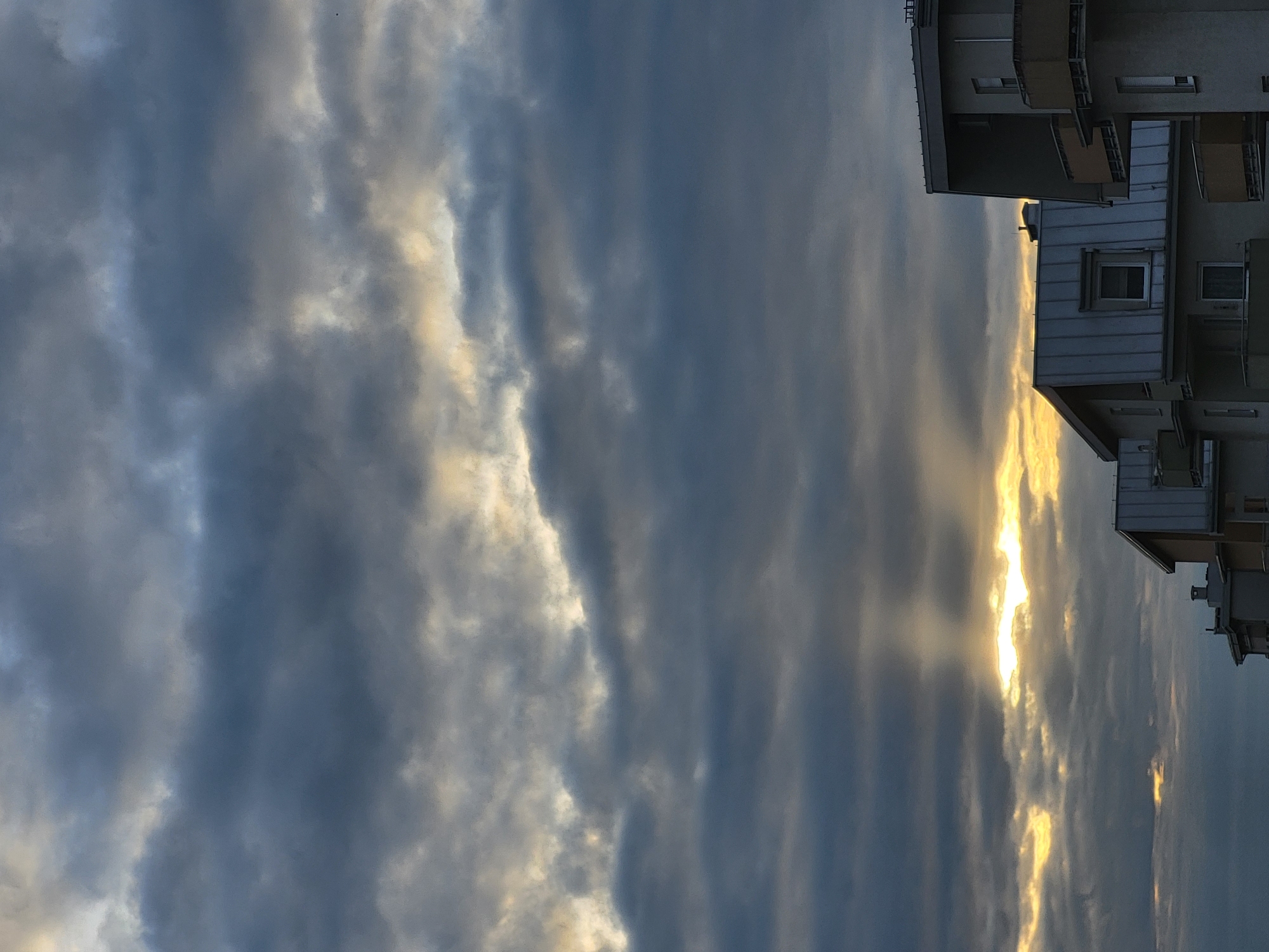 The image shows a dramatic sky with dark, heavy clouds and rays of sunlight breaking through, creating a crepuscular effect. The sun is partially obscured, casting golden light and beams downwards. In the foreground, the rooftops of apartment buildings are visible against the sky. The buildings have modern architecture with flat or sloped roofs, balconies, and visible windows. The overall scene suggests a cloudy day with the sun setting or rising. The location is identified as Unknown, Unknown. There are no people or animals visible in the image.