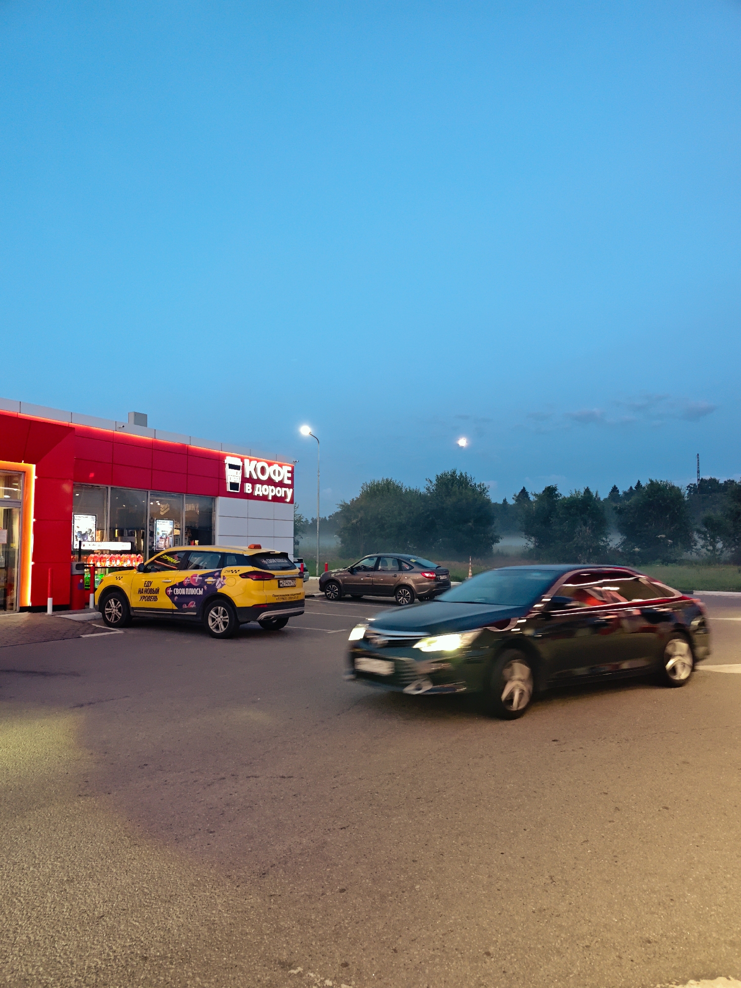 This image captures a scene at dusk outside a red and white building with a sign reading "КОФЕ В ДОРОГУ" (Coffee on the Road), located in Dubrovki, Russia. A yellow taxi is parked to the left of the entrance. To the right, a dark-colored car is in motion, appearing to drive away from the scene. In the background, a line of trees is visible against a pale blue sky with some clouds and a faint moon. The ground is asphalt, suggesting a road or parking area. The lighting indicates it is either early morning or late evening. No people are clearly visible within the frame.