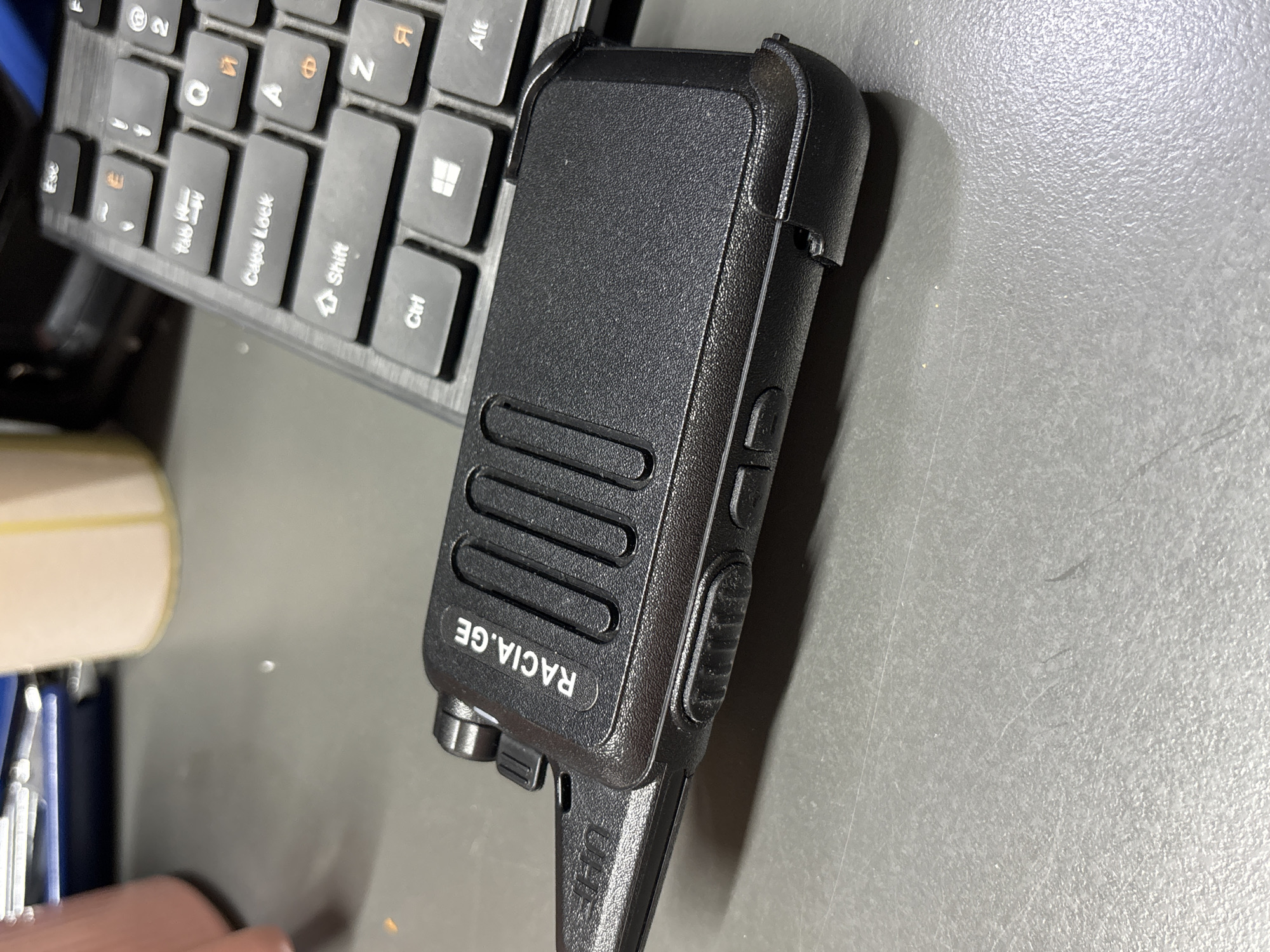 A black two-way radio, labeled "RACIA.GE" and "UHF" is placed on a textured gray surface next to a portion of a black computer keyboard. The keyboard is partially visible, showing keys such as "Esc," "Caps Lock," "Ctrl," "Shift," and several letter keys. The two-way radio is the primary object, with a textured grip and several raised buttons or features on its side. The scene appears to be indoors, likely a workspace, given the presence of the keyboard and the lighting. There are no people visible in the image. The overall impression is one of technology and communication equipment.