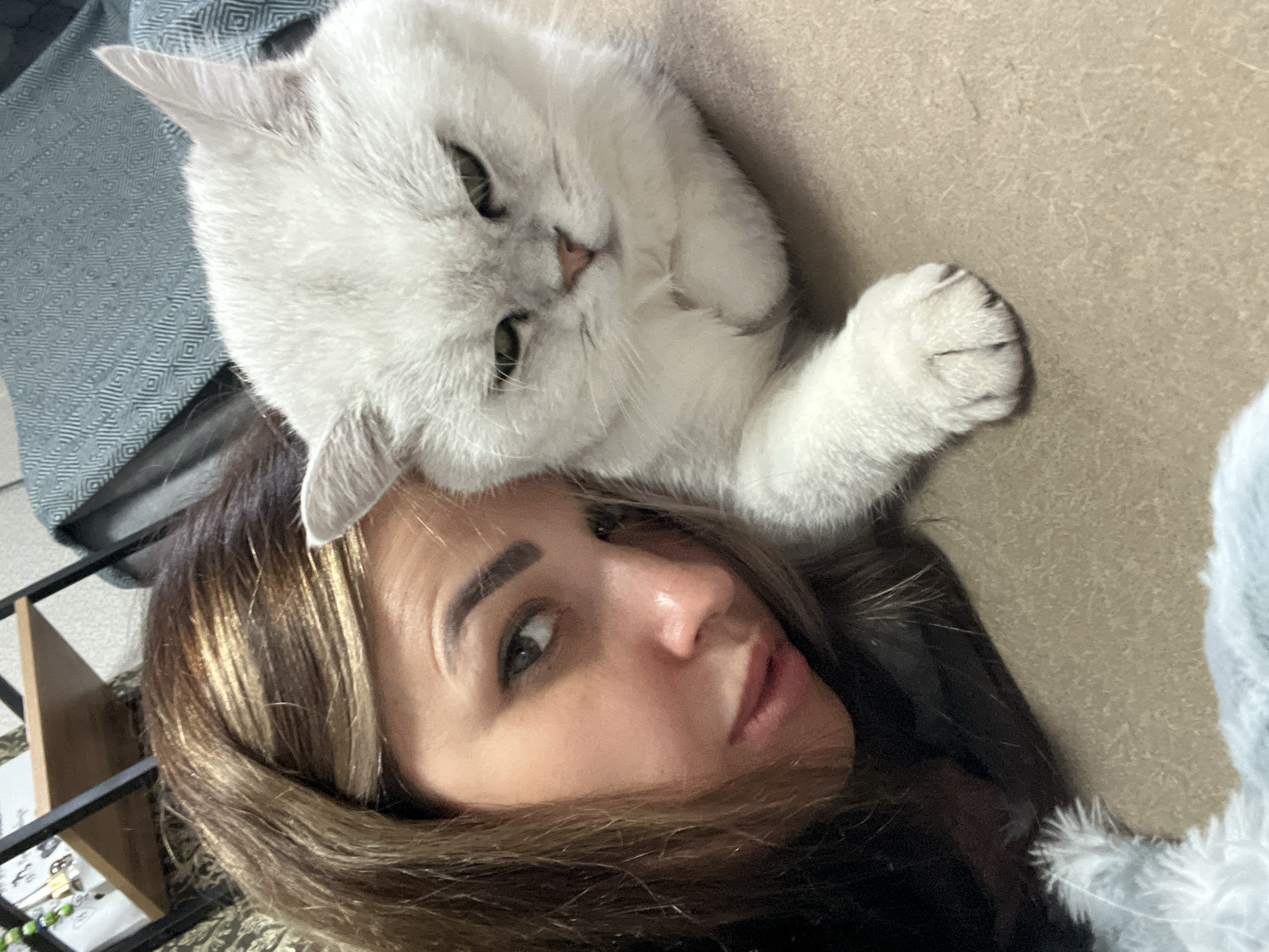 In a close-up, indoor shot, a woman with brown and blonde highlighted hair leans in close to a fluffy white cat with green eyes. The woman's left cheek and eye are visible, along with her nose and lips. The cat is positioned to the right of the woman, facing forward with its head slightly tilted and one paw resting on the surface in front of it. The background is blurred, showing a shelf with objects and a textured blue blanket. The overall tone of the image is intimate and personal, suggesting a moment of affection between the woman and her pet. The location is not specified, and thus cannot be determined to be in any city.