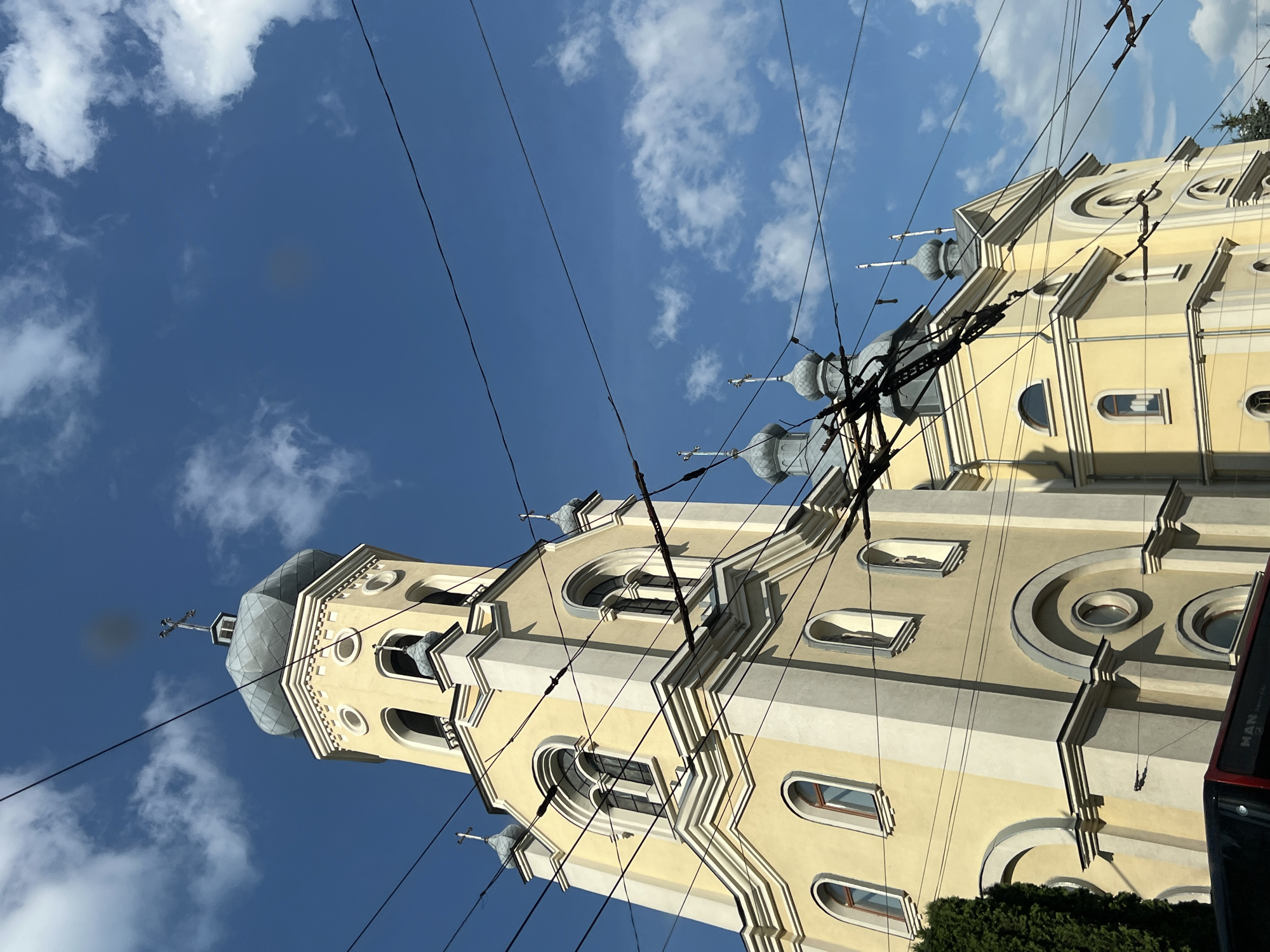 The image shows a yellow church with a tall bell tower and several smaller domes, against a bright blue sky with scattered clouds. Overhead, multiple power lines crisscross the scene, obscuring parts of the church and the sky. The architecture of the church features arched windows and decorative molding. The bottom left of the image shows the top of a red and black bus with the word "MAN" visible. There are no people visible in the frame. This is the Holy Resurrection Cathedral in Ternopil, Ukraine.