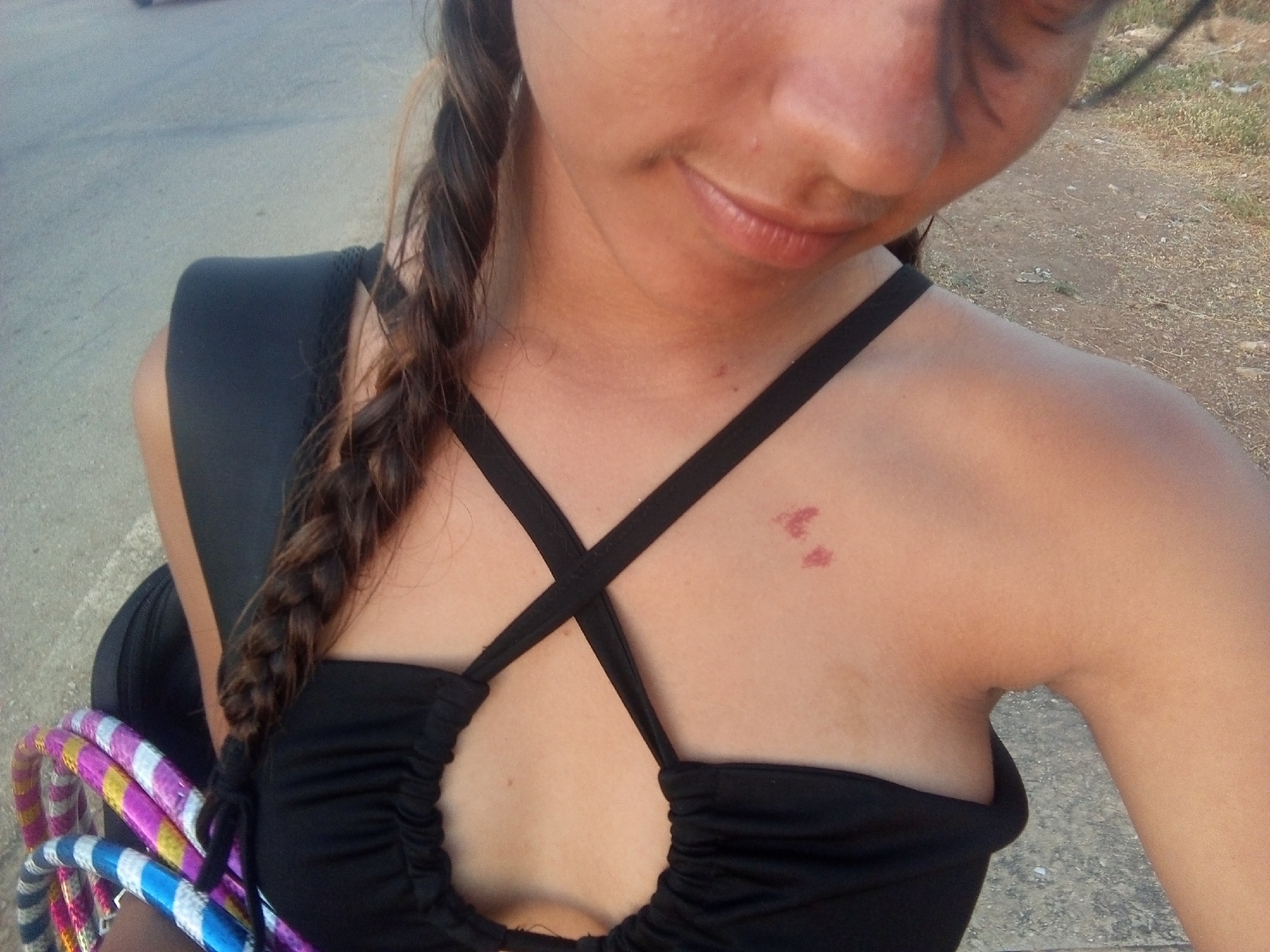 A close-up, slightly angled view shows a light-skinned woman with a brown braid wearing a black top with crisscrossing straps, standing near a street. The woman has a reddish mark on her chest. She carries a black backpack and a collection of colorful hula hoops. The location appears to be outdoors on a street or road adjacent to a gravelly or grassy area. There is no explicit indication or confirmation of the location being Juan Griego, Venezuela in the image.