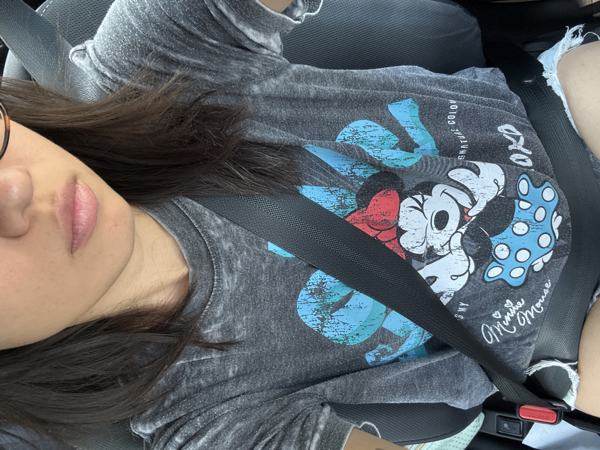 A person wearing a gray t-shirt with a Mickey and Minnie Mouse design is seated in a car and wearing a seatbelt. The person has dark hair and is wearing glasses. They have light skin and their lips are visible. Part of a leg wearing ripped denim shorts is visible on the right side of the image. The interior of the car is mostly dark. There is a "PRESS" button visible on the seatbelt buckle. The specific location is not discernible from the image.