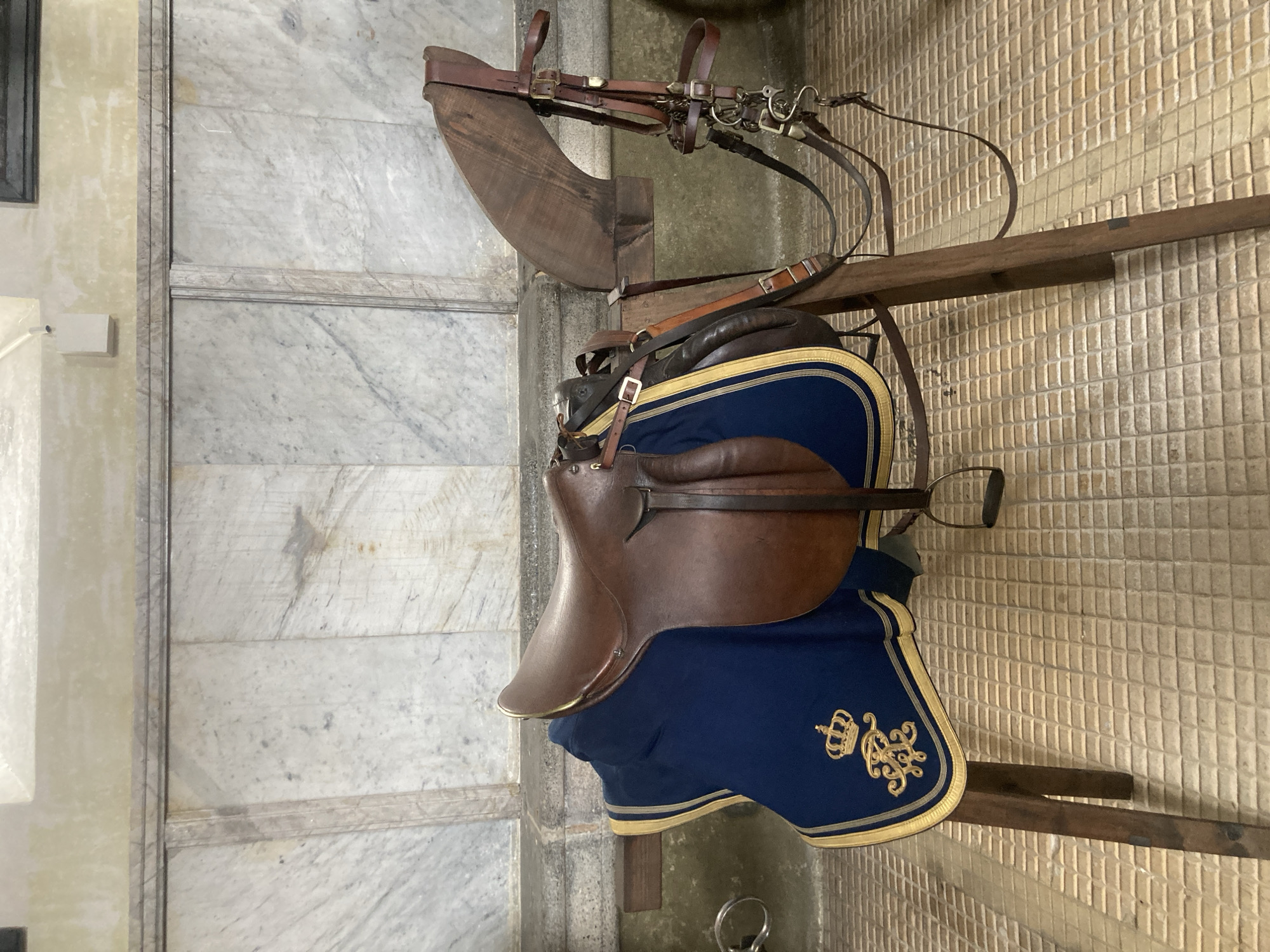 A horse saddle and bridle are displayed on a rack against a wall. The saddle is brown leather with a blue cloth draped underneath, adorned with gold embroidery of a crown and decorative flourishes. The bridle is a complex arrangement of leather straps and metal buckles. The background features two types of wall texture: a white marble-like surface on the left and a tan, small-tiled surface on the right. This is likely a display within a museum or historical site in Sigmaringen, Germany, as the saddle and bridle suggest equestrian history.