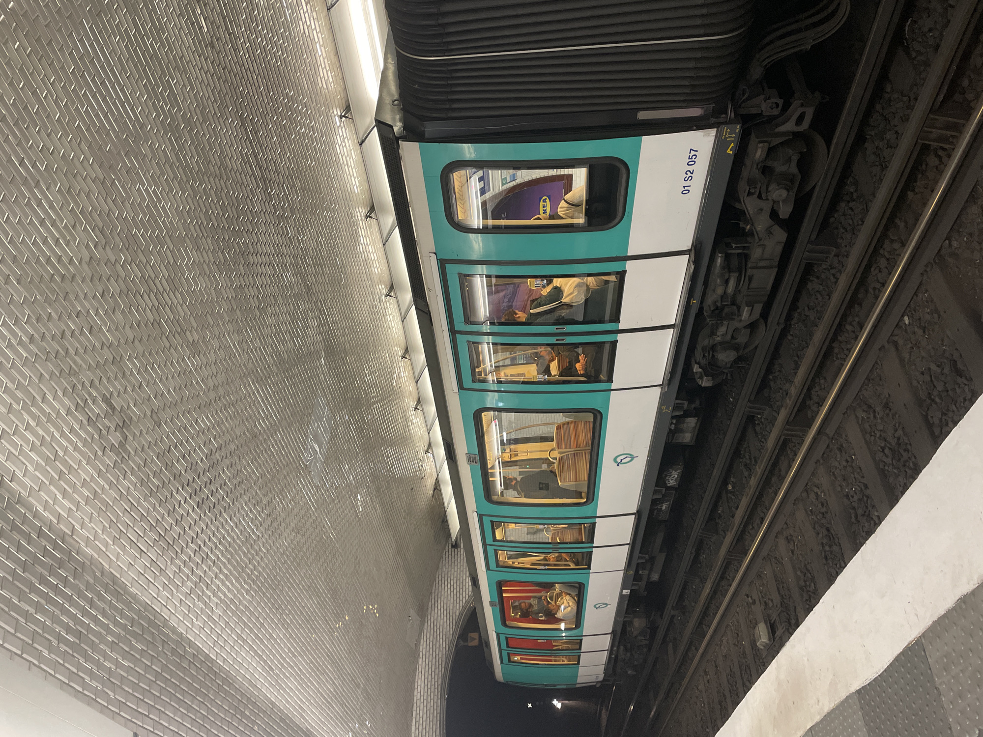 The image shows a train at a subway station. The train is white with a teal stripe and the number "01 S2 057" printed on the side. The interior of the train is visible through the windows, with several passengers seated. The subway station has a curved wall covered in white rectangular tiles. The train tracks are to the right of the train. The location appears to be a Paris Metro station, given the distinctive tiled walls and train design.