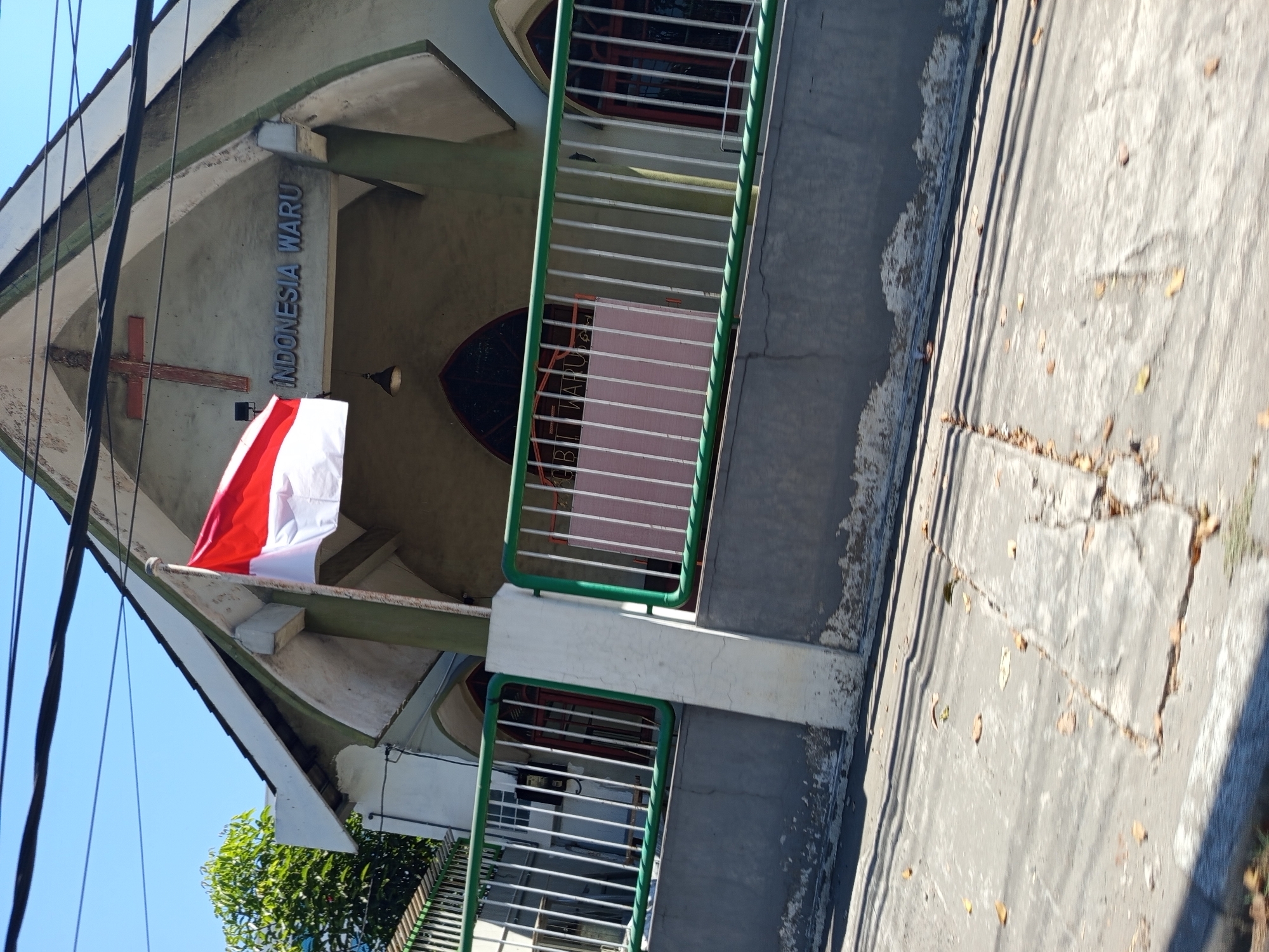 A Dutch colonial era church building with a red cross is seen in Waru, Indonesia. The Indonesian flag flies proudly in front of the building. The building is a light green color with pointed arch windows. The fence in front of the building is green and white. The street in front of the building is cracked and has fallen leaves scattered across it. The sky is a clear blue.