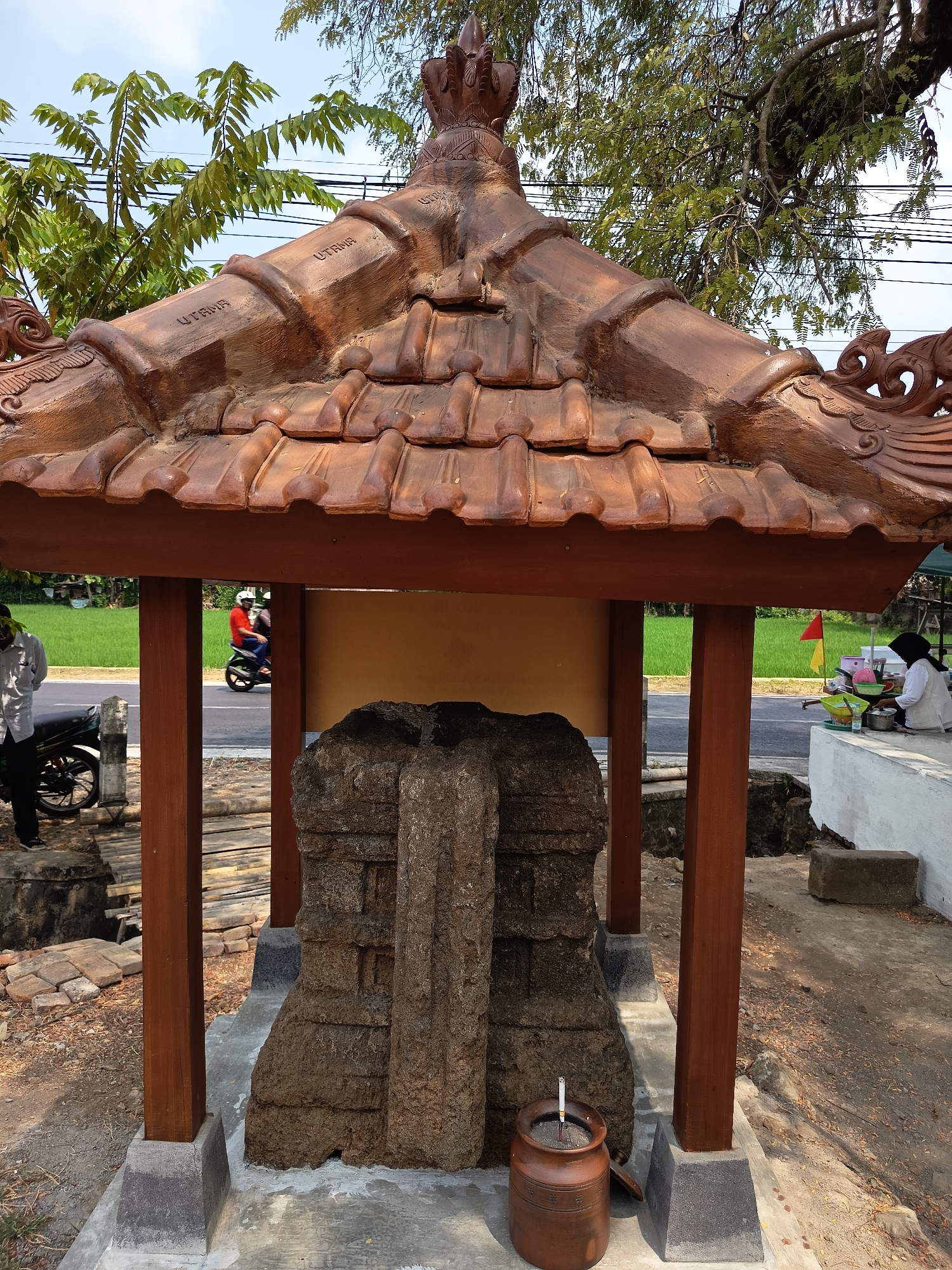 The image depicts a weathered stone artifact housed within a simple wooden structure with a terracotta tiled roof, located outdoors in Lalung, Indonesia. The artifact, which appears to be a monument or ancient relic, is predominantly stone, showing signs of age and erosion with visible carved patterns. The structure protecting it is made of brown wooden posts and beams supporting a brown tiled roof.

In the background, a paved road is visible, with a person on a red motorcycle riding past. To the right, a vendor sits behind a small stall, interacting with a customer. A bright green rice field stretches beyond the road, suggesting a rural or semi-rural setting within Lalung. Overhead, lush green trees with visible branches and leaves contribute to the natural environment, and power lines are present in the upper portion of the frame.

On the left side of the image, another person stands near a parked motorcycle. The ground surrounding the artifact and structure consists of dirt, gravel, and some scattered bricks. A brown ceramic pot filled with sand and a cigarette butt is placed near the base of the stone artifact. The overall scene suggests a roadside attraction or a historical site open to the public.