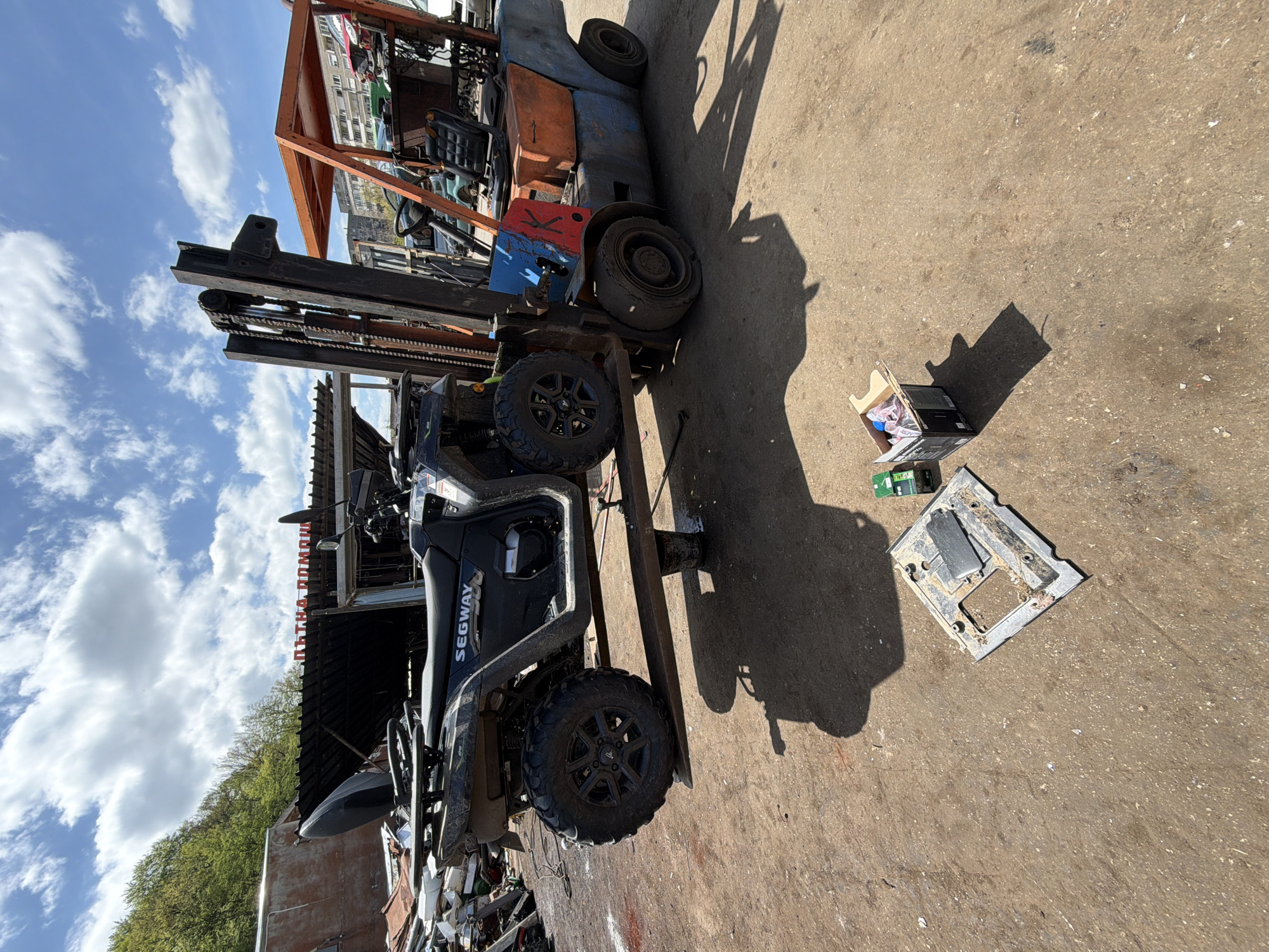 The image shows a scrap yard in Gabrovo, Bulgaria. A Segway ATV is being lifted by an old orange and blue forklift. On the ground, there is a cardboard box with various items inside and a disassembled piece of electronic equipment. The sky is blue with scattered white clouds. The background shows parts of buildings and green trees.