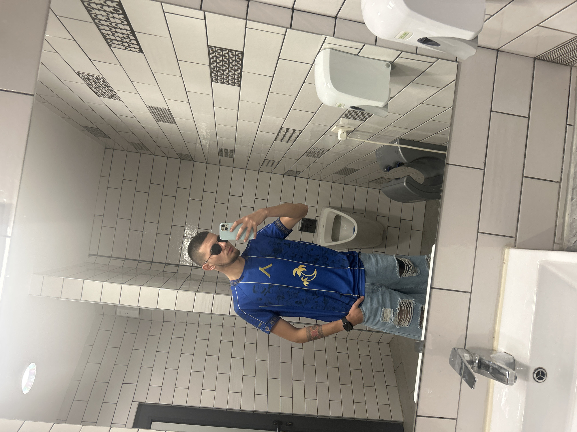 A person is taking a selfie in a public restroom. The restroom has white tile walls with decorative black tile accents. A white toilet, hand dryer, and paper towel dispenser are visible in the mirror's reflection. The person is wearing a blue soccer jersey, ripped jeans, and sunglasses, holding a light blue phone. A sink and faucet are visible on the right side of the frame, with a door at the bottom of the frame. There is no information to confirm the location in the city of Kazanlak, Bulgaria.