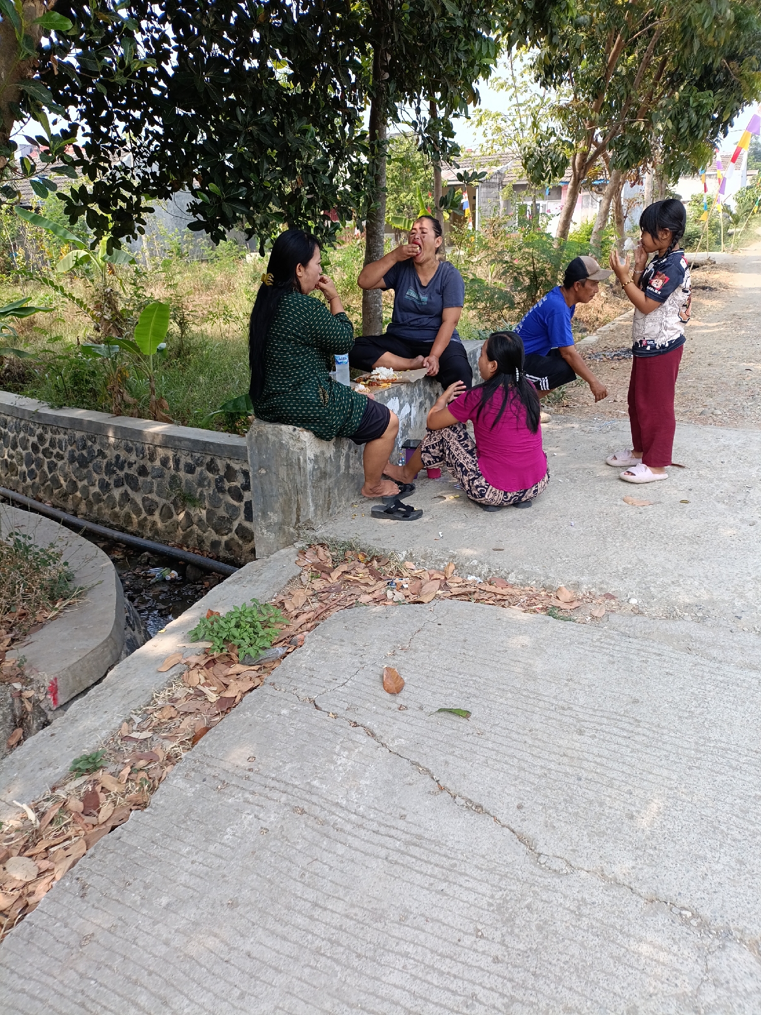 In Cariu, Indonesia, on a sunny day, a group of five individuals, including two adults and three children, are gathered outdoors near a concrete structure adjacent to a small drainage ditch. One adult woman, wearing a green patterned top and dark shorts, sits on the edge of the concrete structure, her back to the camera, and appears to be eating. Another adult woman, dressed in a dark t-shirt and dark pants, sits to her left, also seemingly engaged in eating and with her mouth open. A man wearing a blue shirt and a baseball cap is crouched to the right of the seated women, facing towards the children. Two children are present: one girl in a colorful t-shirt and maroon pants stands to the right, holding a clear plastic cup and looking towards the man, while another girl in a pink shirt and patterned pants squats on the ground to the left, facing the seated women. A white platter with food is visible on the concrete in front of the seated women. The environment includes trees with green foliage overhead, a stone wall lining the drainage ditch, and a dirt path or road in the background. Flags are also visible in the distance. The ground is concrete, with scattered dry leaves and some greenery.