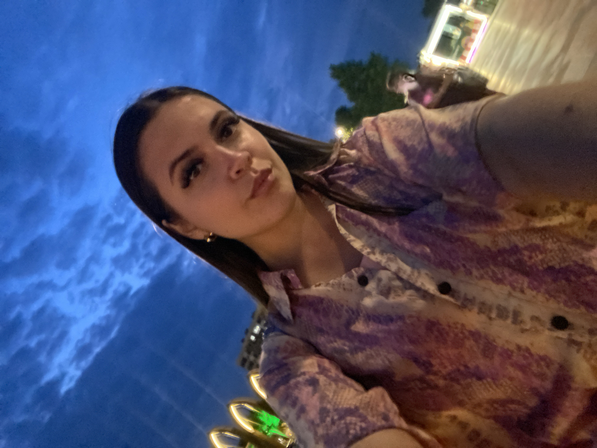 A young woman with long dark hair and full makeup is taking a selfie outdoors at dusk. She is wearing a long-sleeved button-up shirt with a pink and purple snakeskin pattern. The background shows a deep blue sky with scattered clouds, illuminated trees, and blurred lights of buildings and possibly an amusement park. The location is the city of Derbent, Russia.
