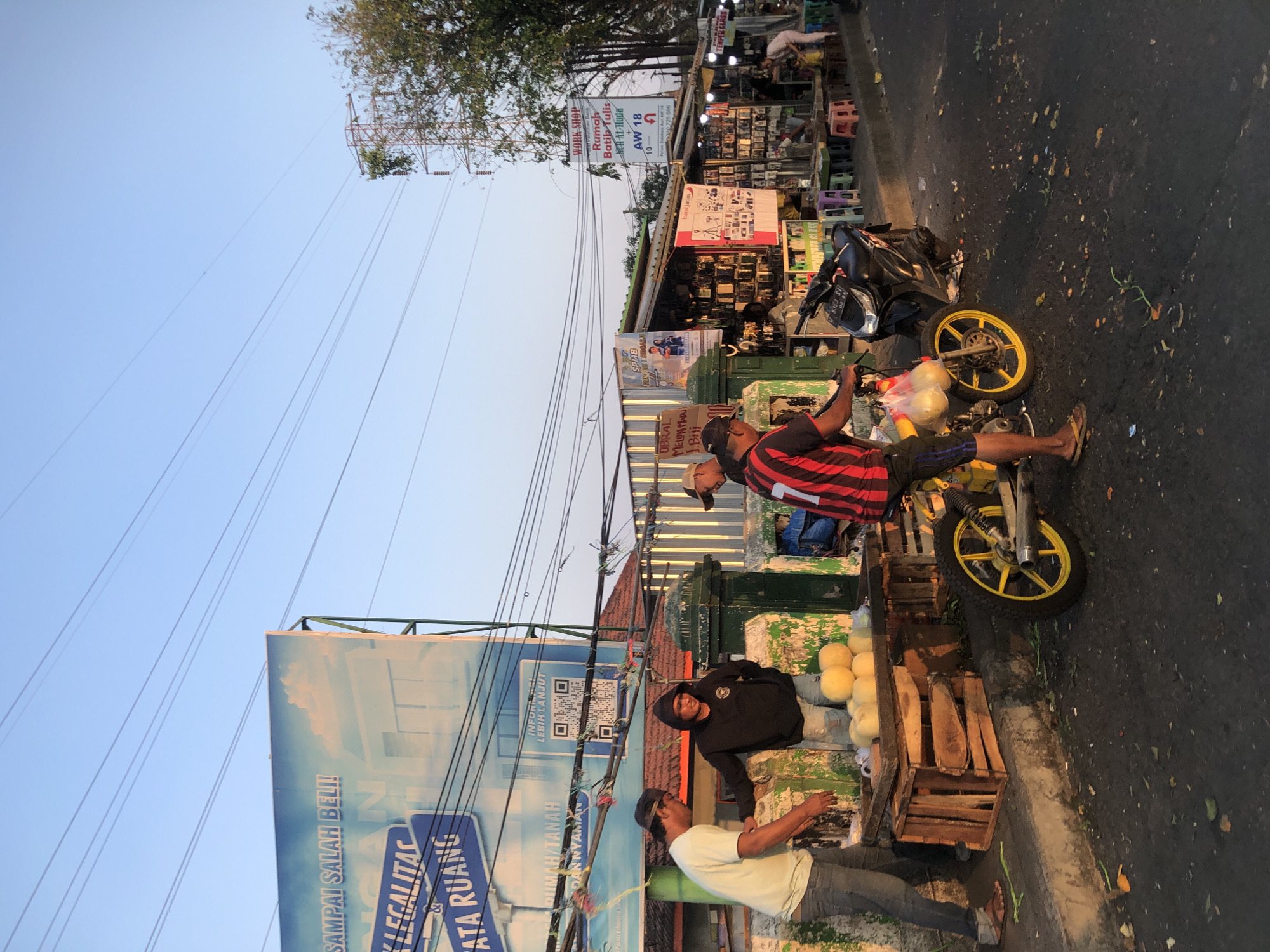 The image depicts a street scene in Sepande, Indonesia, at what appears to be late afternoon, given the low angle of the sunlight. The foreground is dominated by a street with scattered debris, including green leaves and orange peelings. A motorcycle with yellow wheels is parked on the right side, and a person wearing a black and red striped shirt and blue shorts is standing next to it, holding a plastic bag with some round yellow fruits.
To the left of the motorcycle, a wooden cart is filled with several round yellow fruits, possibly melons. Three men are gathered around the cart. One man in a white t-shirt and jeans is facing away from the camera, appearing to interact with the fruits. Another man in a black hoodie and ripped jeans is looking down at the fruits. A third man, wearing a black cap and a black and red striped shirt, is partially visible on the far right.
Behind the cart, a green painted structure with a mossy texture is visible, serving as a wall or barrier. Above this, a large blue billboard with white text is displayed, featuring images of buildings and clouds. Several power lines crisscross the sky, supported by utility poles, one of which is prominent on the right side of the image.
Further down the street, a row of small shops or stalls are lined up, selling various goods. Signs are visible above these stalls, including one advertising "WORK SHOP" and another with text in Indonesian. People are visible in front of these shops, suggesting ongoing commercial activity. The overall impression is of a busy local marketplace or street vendor area.