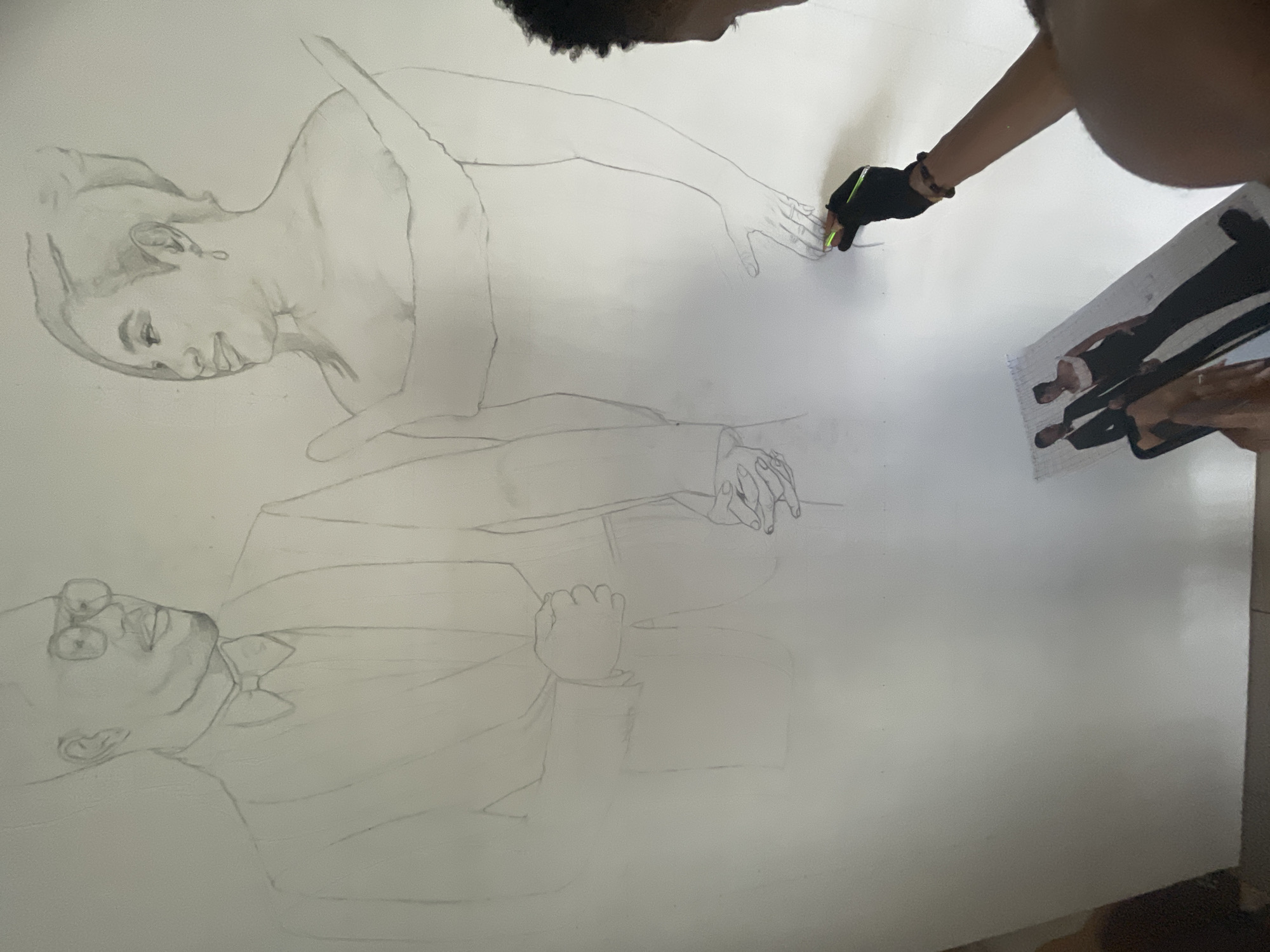 The image depicts a person drawing a portrait on a large white canvas. The portrait features two figures, a woman and a man, rendered in pencil. The woman is shown in the upper portion of the canvas. The man, positioned below, wears glasses and a suit. The person drawing is visible in the top right corner, using a pencil with their right hand, wearing a black glove. A reference photograph lies beside the canvas, showing the full figures of the same couple. The scene is indoors, likely in a studio or workspace. It is unknown if the location is in Ado, Nigeria.