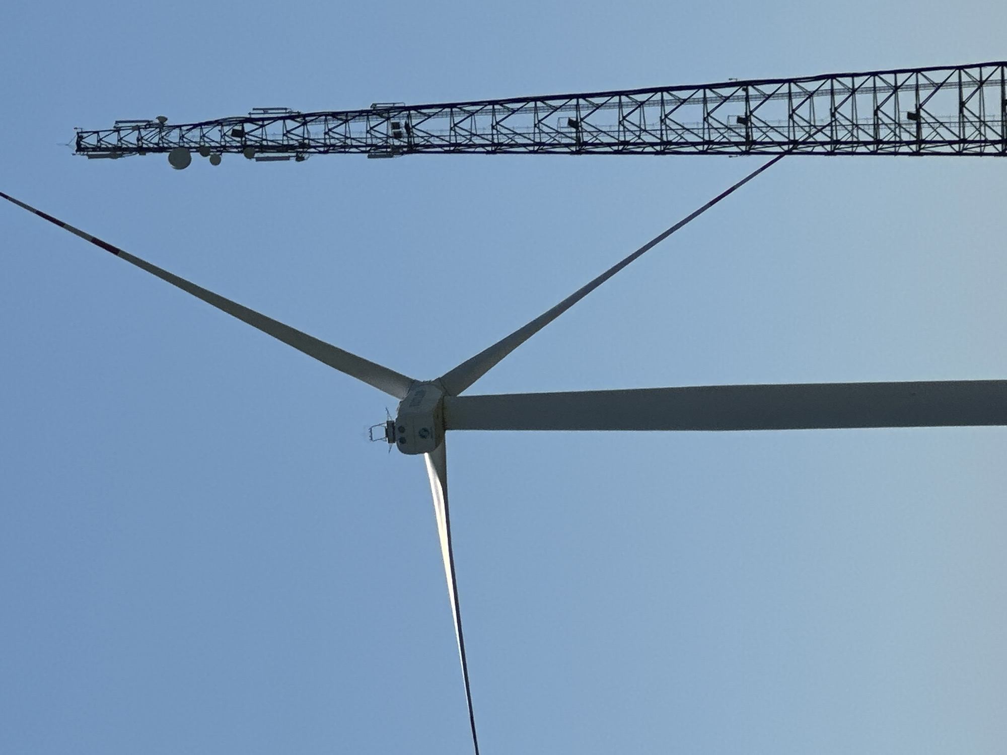 An image shows a clear blue sky behind a wind turbine and a telecommunications tower. The wind turbine is on the left side of the frame, and its white blades are extended. The telecommunications tower is on the right side of the frame, with a dark, lattice-like structure and several antennas. The scene is likely an outdoor landscape, possibly in a rural or industrial area given the presence of these structures.