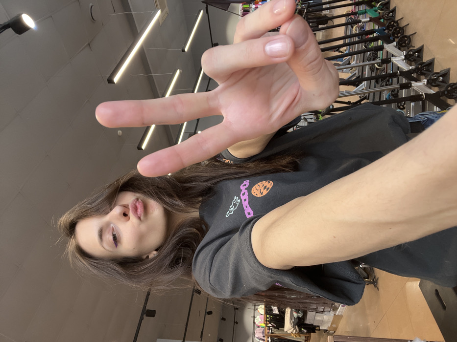 The scene depicts a young adult female posing for a selfie inside a retail store, located in the city of Krasnoyarsk, Russia. The woman has long, dark, slightly disheveled hair, and is wearing a dark-colored oversized t-shirt with a multi-colored graphic print featuring an orange circle and purple/pink abstract shapes. She has purple eyeliner visible on her left eye and a small ear piercing. Her lips are pursed in a pouting expression, and her right hand is raised towards the camera, forming a "V" (peace) sign with her extended index and middle fingers. The background reveals a modern commercial interior with a light-colored ceiling featuring black track lighting fixtures and linear LED lights. To the immediate right of the subject, a prominent display of multiple kick-scooters or stunt scooters is visible, arranged in organized rows, suggesting a specialized sports or leisure goods store. Blurry shelving with other merchandise is faintly discernible in the background to the left.