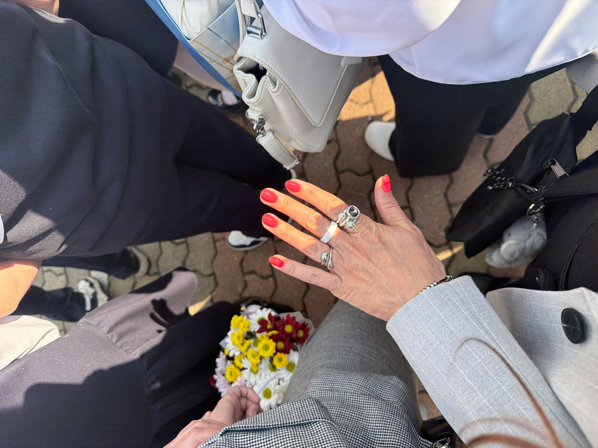 A high-angle shot shows a group of people standing close together, seemingly outdoors. A woman's hand, prominently displaying red nail polish and several rings, is centrally placed. She is holding a bouquet of flowers consisting of yellow, red, and white daisies. The person is wearing a grey jacket with pinstripes, and pants with a checkered pattern. The location is not possible to identify.