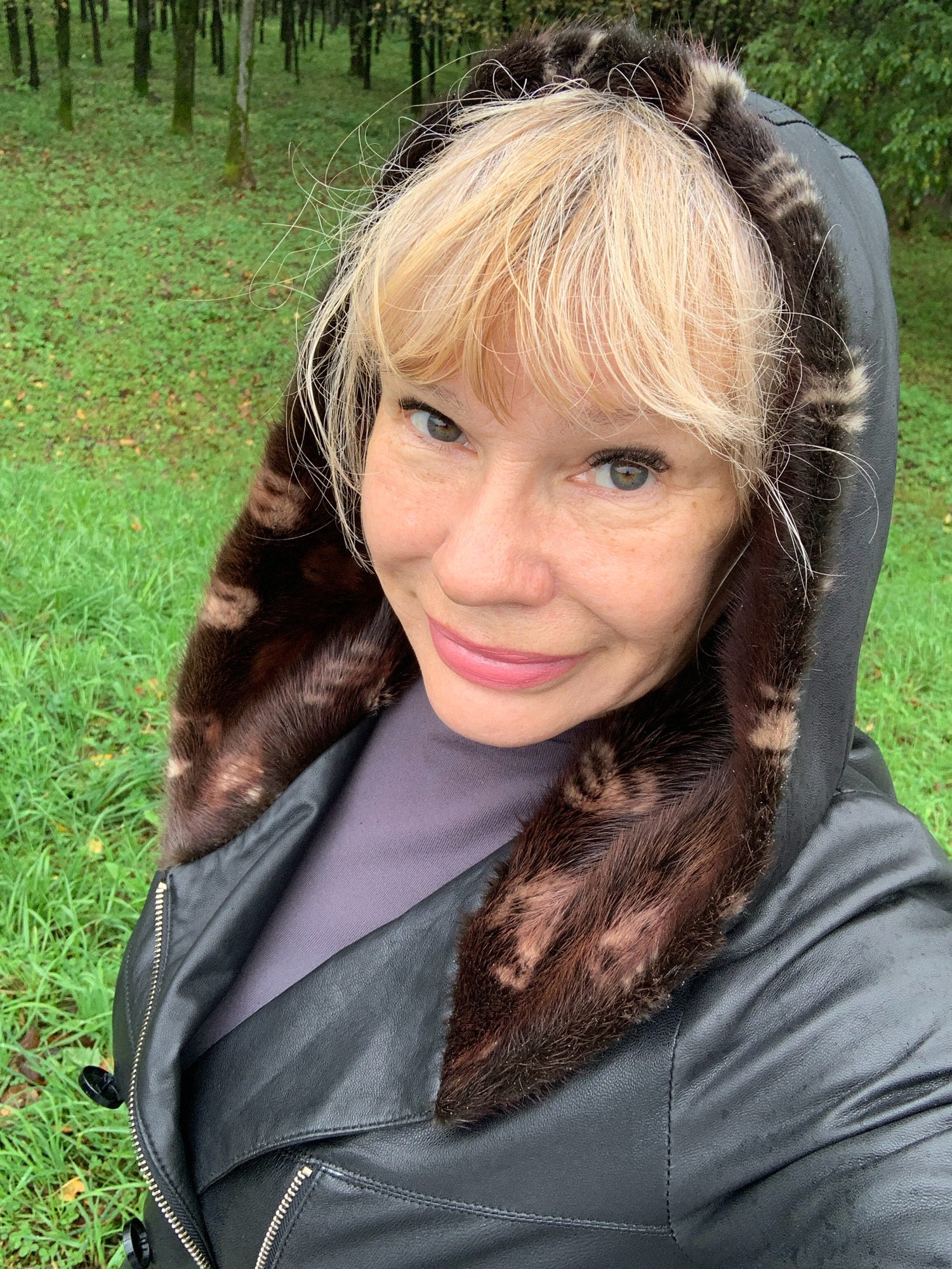 A middle-aged Caucasian woman with blonde hair and light skin is posing for a selfie outdoors. She is wearing a black leather jacket with a fur-lined hood and a dark purple shirt underneath. The background is a grassy area with trees, suggesting a park or forest setting. There are no specific landmarks visible to identify the location in Prokopyevsk, Russia. The woman is smiling and looking at the camera.
