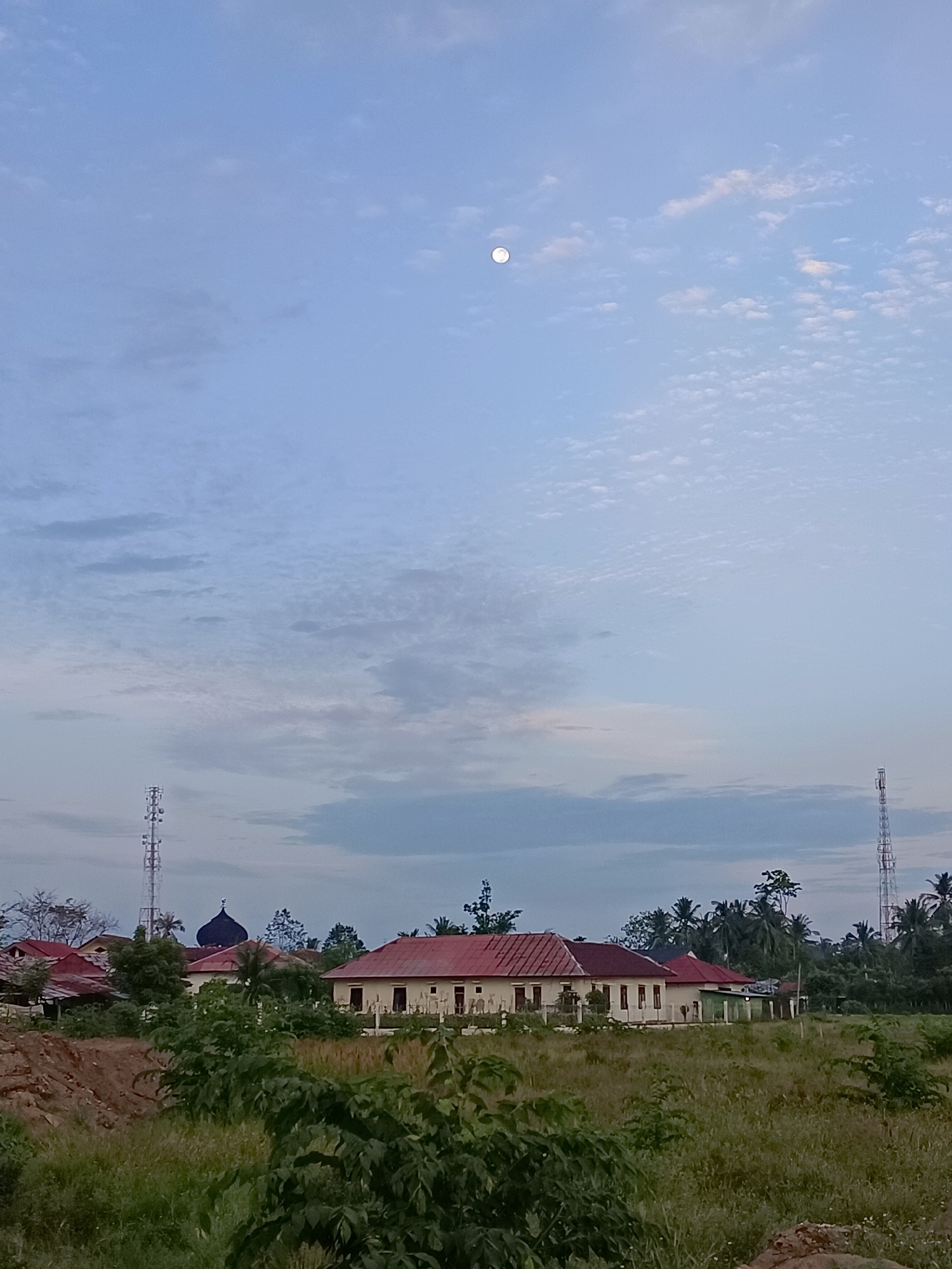The image depicts a tranquil scene in Meunasah Pupu, Indonesia. The sky is a pale blue, dotted with wispy clouds, and a full moon is visible in the upper portion of the frame. In the middle ground, a long, single-story building with a red-tiled roof stands prominently. To the left of this building, a dark, domed structure, likely a mosque, is partially visible. Two tall telecommunication towers flank the scene, one on the left and another on the right, reaching towards the sky. The foreground is filled with lush green vegetation, suggesting a rural or semi-rural environment. The overall impression is one of a peaceful evening in a Indonesian village.