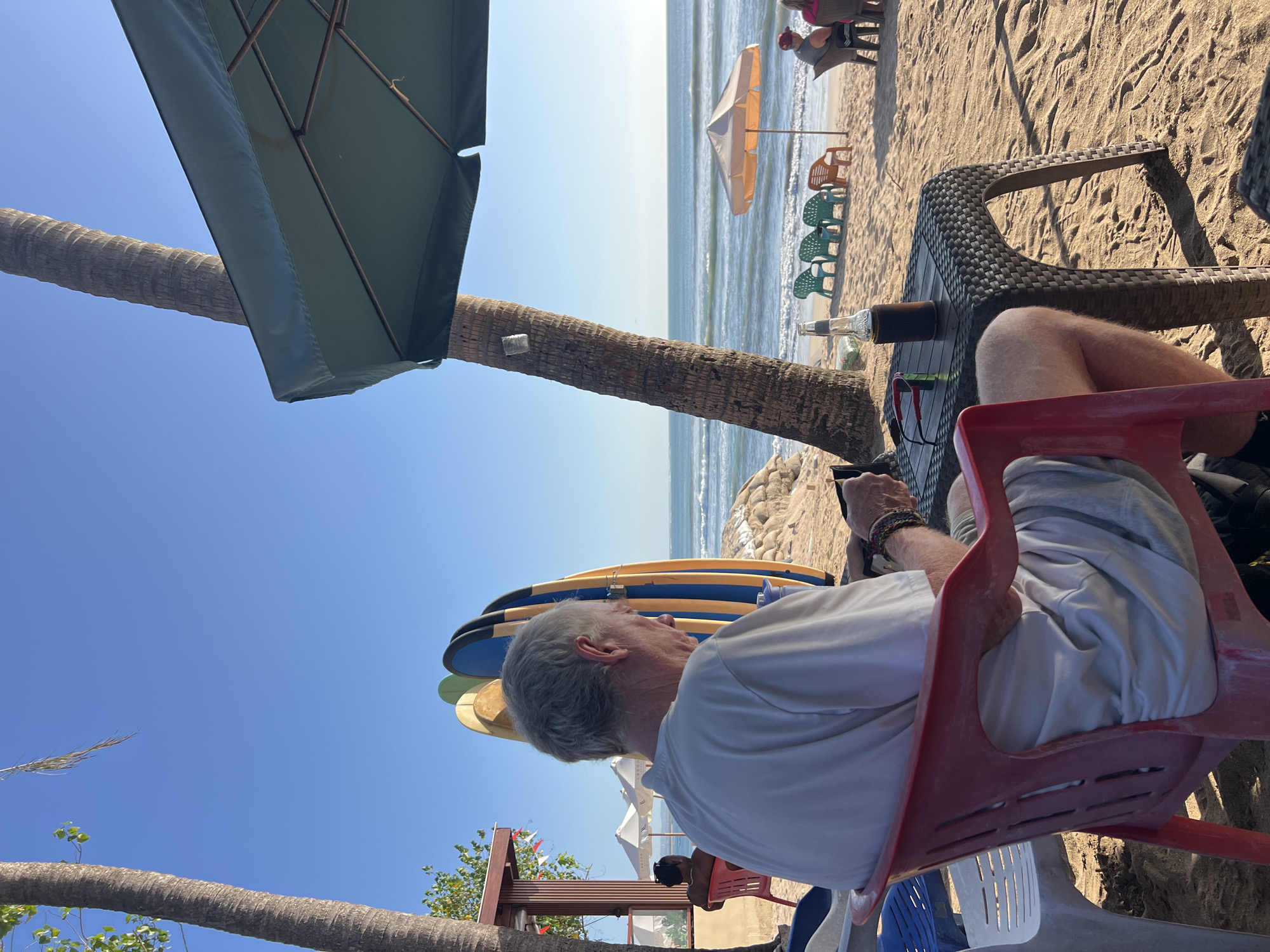 In Kuta, Indonesia, an elderly man with gray hair sits at a textured plastic table on a sandy beach, facing away from the camera. He is wearing a light-colored t-shirt and shorts. In front of him on the table are a bottle of drink in a cooler, a pair of sunglasses, and a phone. The beach is lined with palm trees, surfboards, and lounge chairs. In the background, the ocean meets a clear blue sky. Other people are visible further down the beach, seated at tables and under umbrellas.