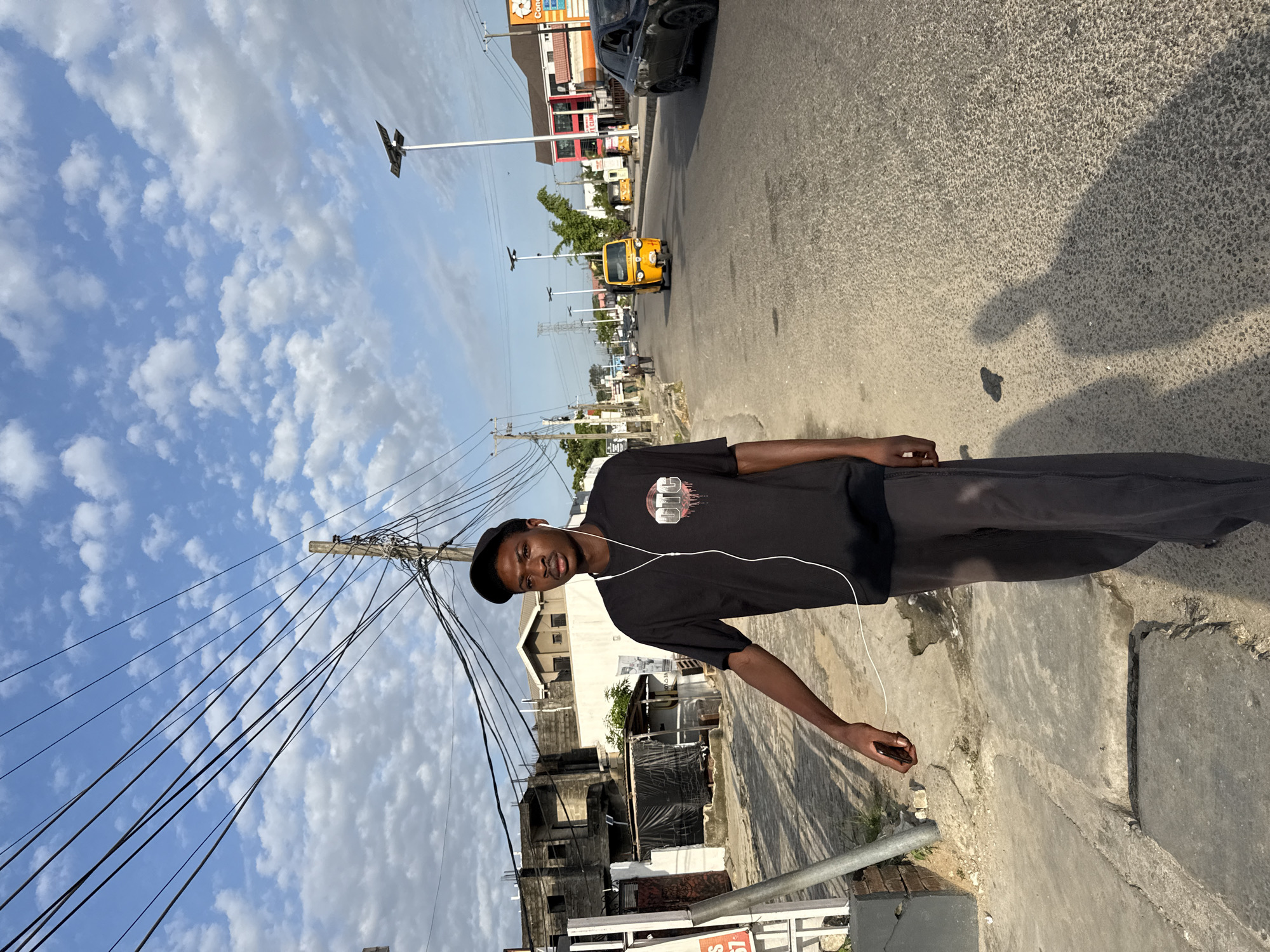A young man stands on a sidewalk in Ado, Nigeria. He wears a black t-shirt, dark pants, and a cap. He has earphones on and appears to be holding something small in his right hand. Behind him is a street with a yellow tricycle vehicle, cars, and buildings. A pole with a multitude of wires runs along the edge of the sidewalk. The sky is blue with scattered clouds. The image appears to be taken in daylight.