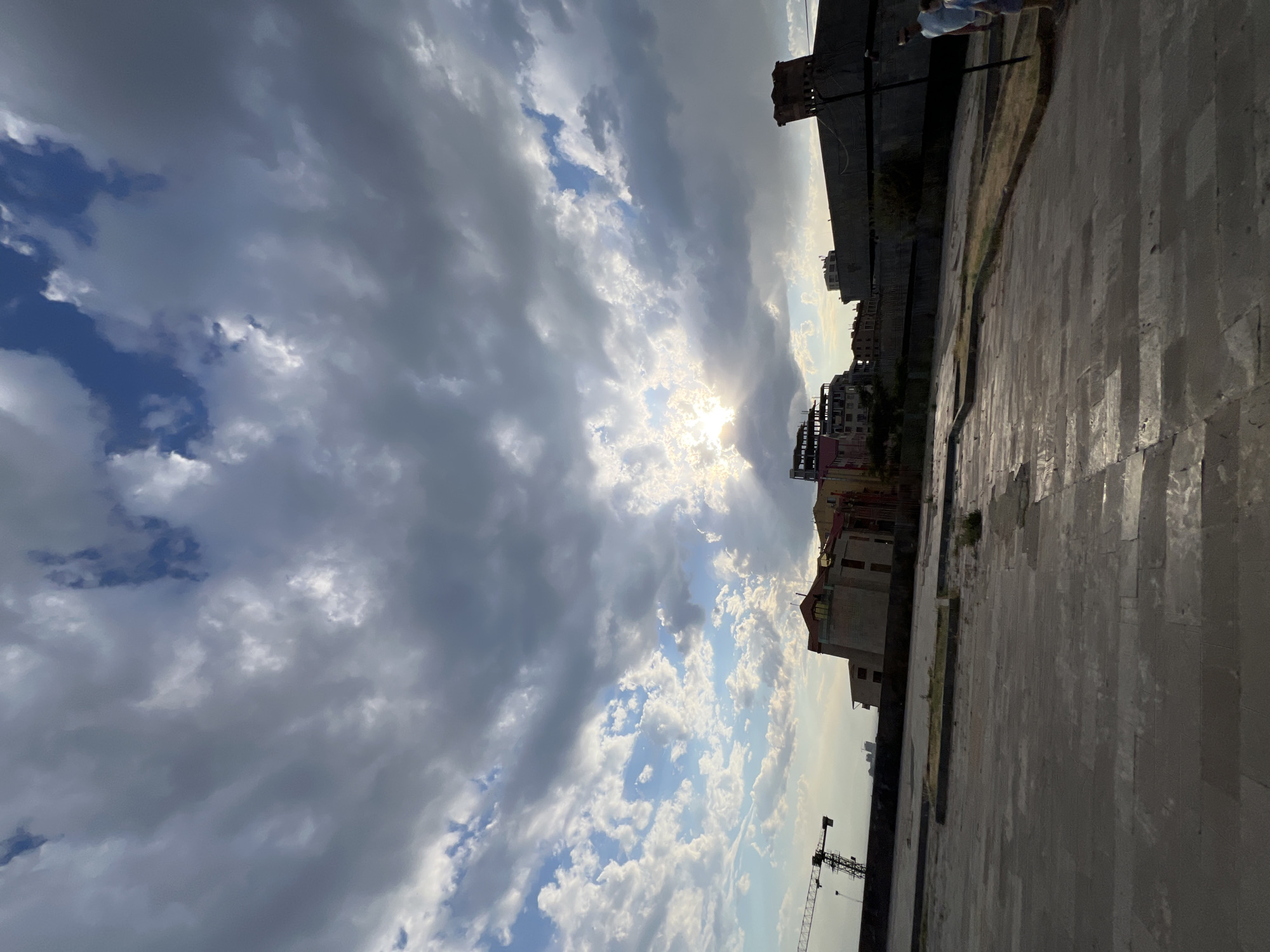The image depicts an outdoor scene on a cloudy day, possibly in Yerevan, Armenia, given the context. The sun is peeking through the clouds, creating a dramatic lighting effect with bright rays and shadows. The sky is filled with a mix of dark and light clouds, with patches of blue visible.
In the foreground, there is a paved area made of stone tiles, which appears to be a public space or plaza. The ground is illuminated by the sunlight, casting long shadows.
Along the edge of the paved area, there are several buildings in various stages of construction or renovation. Some appear to be older, multi-story structures with distinct architectural features, while others look like modern constructions with scaffolding. A construction crane is visible in the background, further indicating ongoing development.
A few people can be seen in the distance, near the top of the image, their figures small against the vastness of the sky and cityscape. They are indistinct but suggest human presence in the area.
The overall atmosphere is somewhat overcast due to the clouds, but the bright sunlight breaking through adds a touch of hope and dynamism to the scene. The time of day appears to be late afternoon or early evening, given the angle of the sun and the long shadows. There is no visible text in the image.