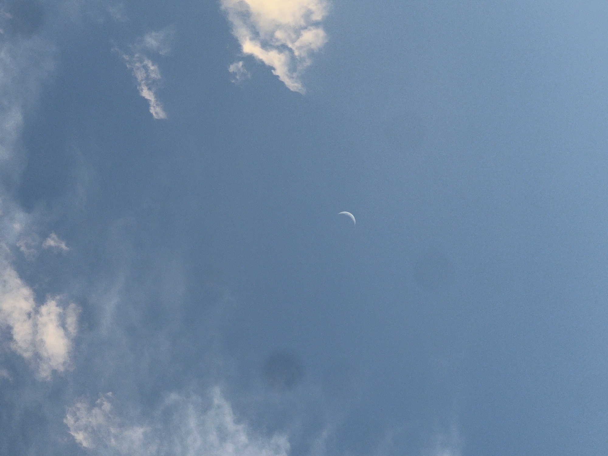 The image shows a clear blue sky with scattered white clouds. A crescent moon is visible in the upper right quadrant of the sky. There is no indication of the city of Batumi, Georgia, in the image.