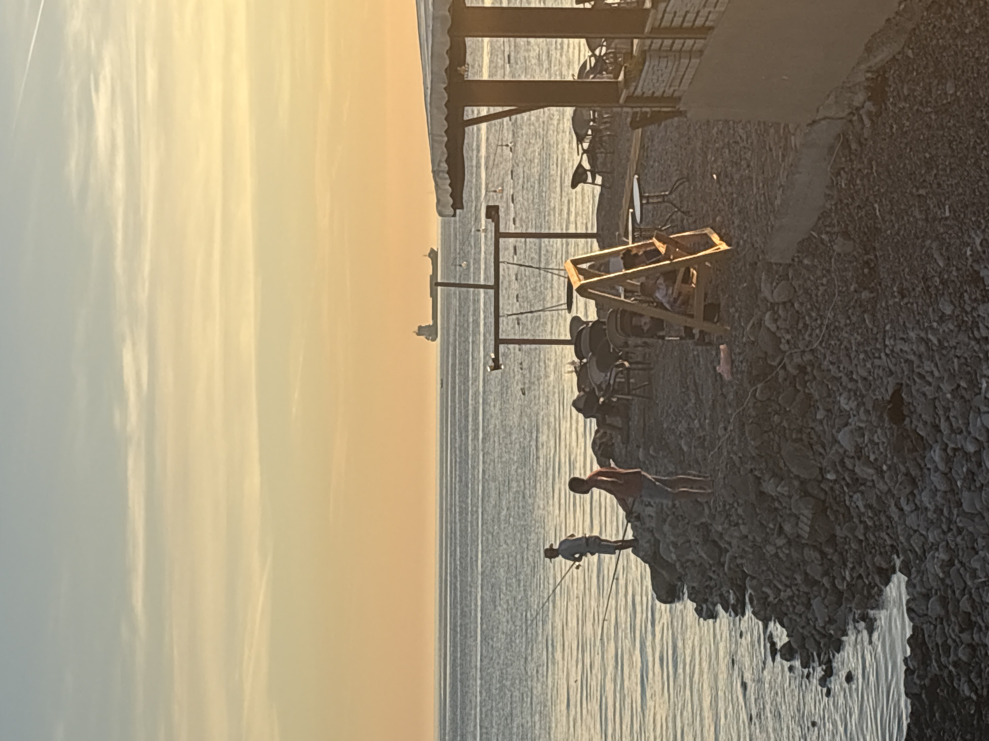 The image depicts a scene on a beach in Bar, Montenegro, during either sunrise or sunset. Two individuals are fishing from a rocky outcrop by the sea. The water is calm, and a large cargo ship is visible on the horizon. To the right of the image, there is a covered seating area with tables and chairs, suggesting a cafe or restaurant. Further inland, a swing set is present. The sky is a gradient of soft yellow and blue, indicating the time of day. The overall atmosphere is peaceful and serene.
