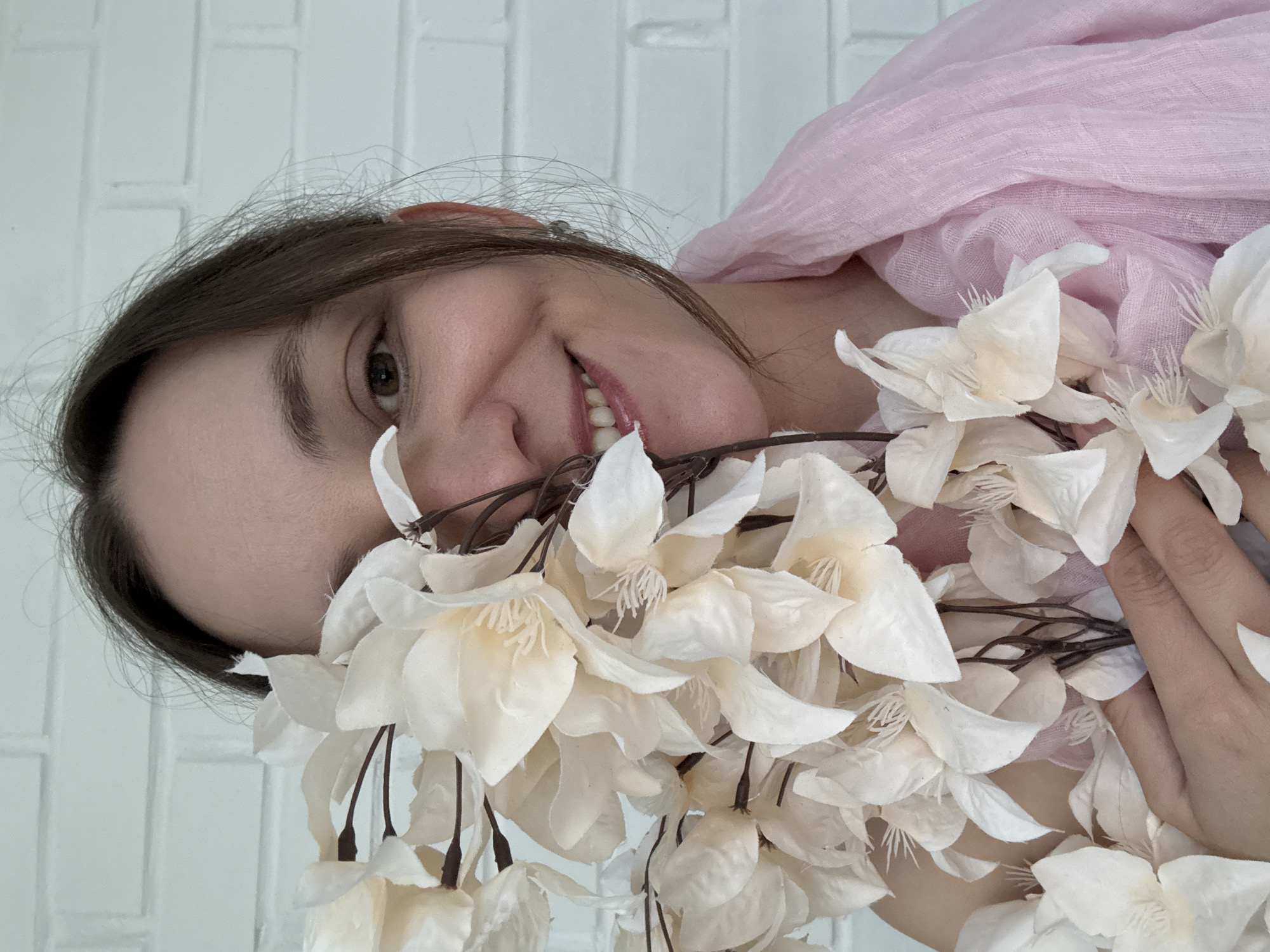 The image, oriented vertically with a clockwise rotation, features a person, likely a woman, with fair skin, dark brown hair, and brown eyes, smiling directly at the camera. Her head is tilted, and her expression is gentle and happy, with her mouth slightly open to reveal white teeth.
She is partially draped in a light pink, sheer fabric, possibly a scarf or shawl, visible over her upper body on the right side of the frame. Her right hand is visible on the far right, holding a large cluster of artificial white or off-white flowers with dark brown stems. These fabric flowers, resembling delicate blossoms with prominent stamens, occupy a significant portion of the lower and central image, partially framing her face.
The background consists of a uniformly white brick wall, suggesting an indoor or sheltered setting with a clean and bright aesthetic. The lighting is soft and even, highlighting the subject's features and the textures of the flowers and fabric. No text is visible in the image. The overall mood is serene and pleasant.