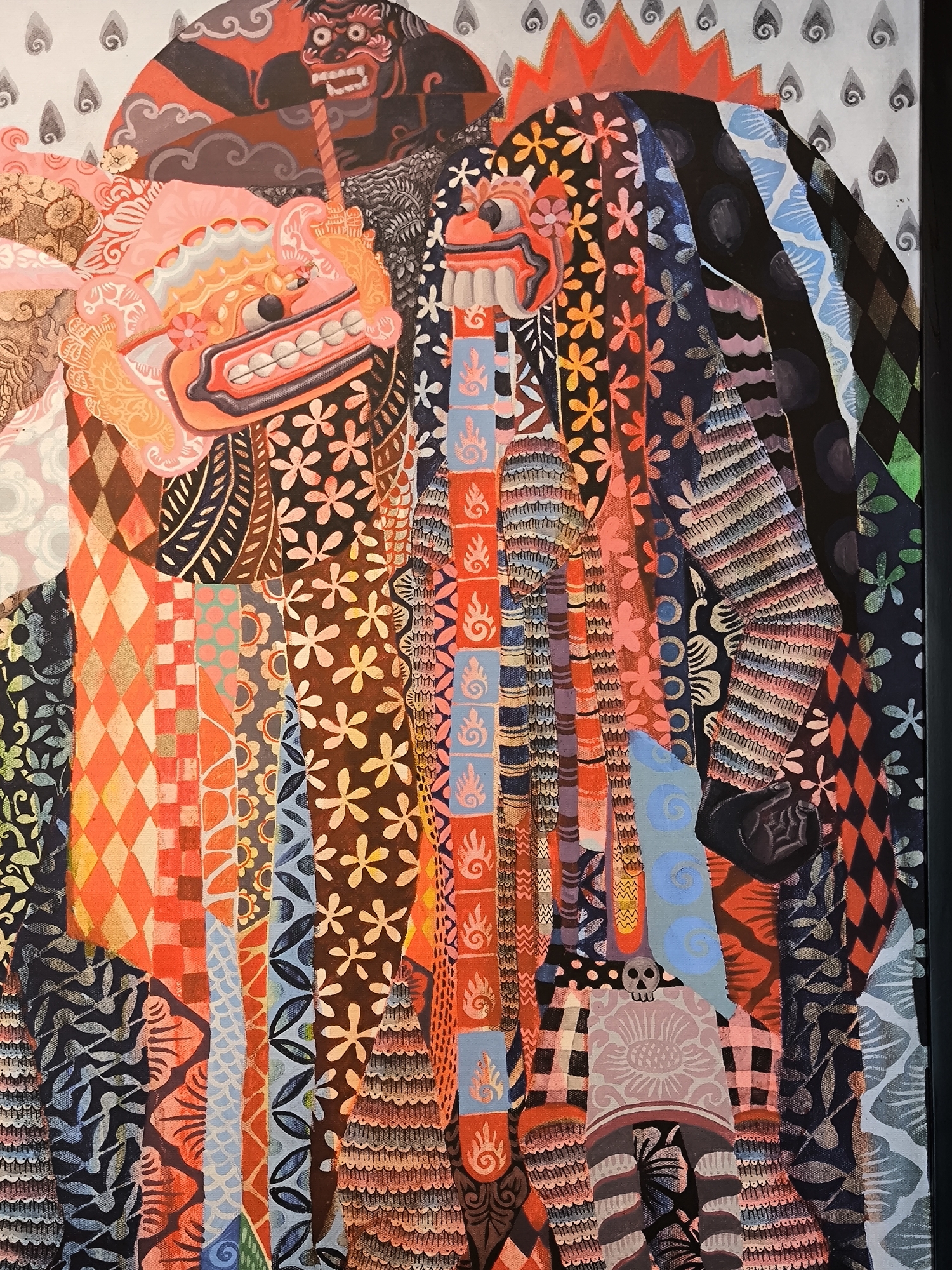 The image is a close-up of a colorful, stylized painting featuring depictions of Balinese Barong masks. The masks are adorned with intricate patterns, floral motifs, and geometric designs. Various colors are used, including shades of red, orange, brown, blue, black, and pink. The background includes abstract shapes and patterns. The overall style suggests a folk art or traditional craft aesthetic.
