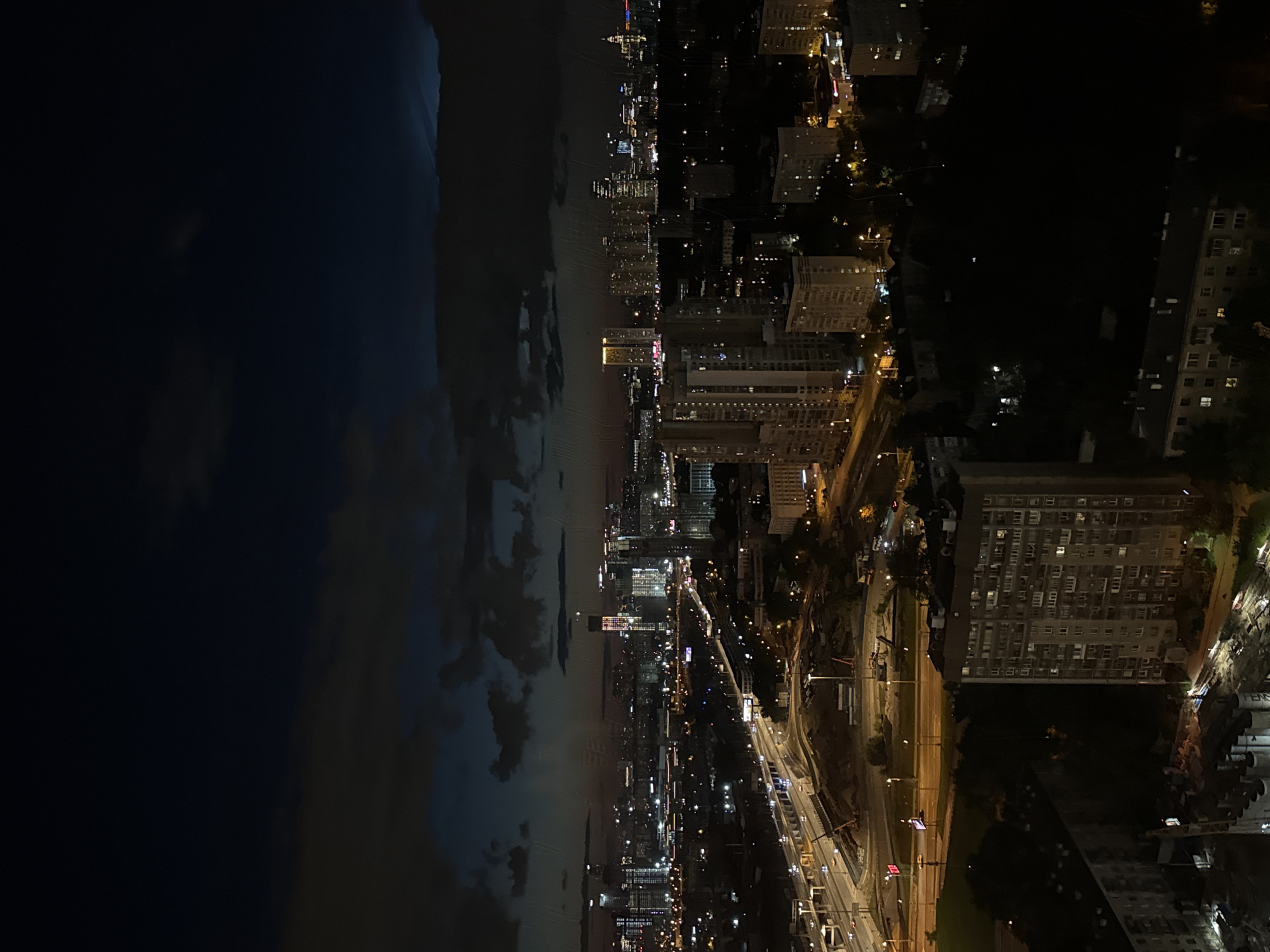 This media file is a nighttime, high-angle view of a city, likely Khimki, Russia, showcasing a blend of urban landscapes and natural elements. The scene is divided by a dark, expansive body of water on the left, reflecting the faint lights of the city across it. Dense, dark clouds loom over the water, creating a dramatic contrast with the illuminated urban sprawl on the right.
The city is characterized by numerous buildings of varying heights, many of which are brightly lit, indicating nighttime activity. A prominent feature is a multi-lane highway with flowing traffic, its headlights and taillights creating streaks of light that snake through the cityscape. Bridges and elevated roadways are also visible, contributing to the complex infrastructure. In the foreground, closer to the viewer, are several large, apartment-style buildings with numerous illuminated windows. Trees are sparsely scattered throughout the urban area, their forms silhouetted against the lights.
The time of day is clearly night, with the sky dominated by darkness and clouds, and the city illuminated by artificial lights. The weather appears to be somewhat overcast, contributing to the dark and moody atmosphere. The overall impression is one of a bustling city at night, observed from a distance, with the imposing presence of the water and clouds adding a natural, almost atmospheric, dimension to the urban scene. There is no visible text in the image.