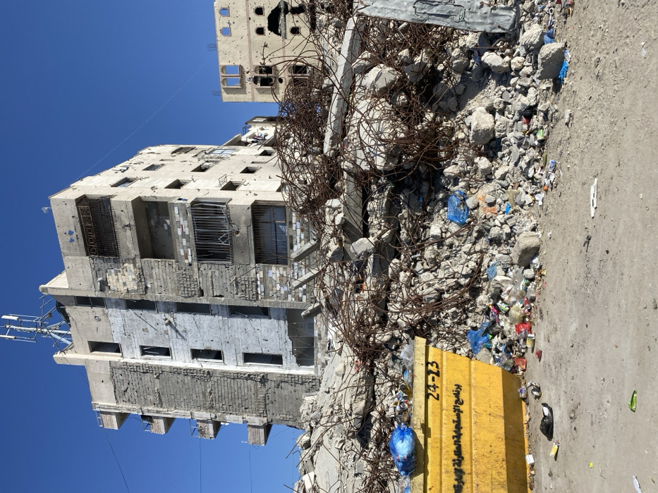 The image captures a scene of extensive urban destruction under a clear, bright blue sky, suggesting daytime. The central focus is a multi-story concrete building on the left side, which appears heavily damaged. Its facade is pockmarked with numerous small holes, consistent with bullet or shrapnel impacts. Many windows are shattered, missing glass, or feature bent and twisted metal grates. Some balconies are visibly damaged or partially collapsed, and exposed rebar protrudes from broken sections of concrete. The lower levels of this building show varying textures and shades of grey and white, indicating repairs or different construction materials, all bearing signs of wear and damage. An antenna or communication structure is visible on the upper part of this building.
Behind the prominent damaged building, another concrete structure is partially visible in the background, also showing similar signs of damage, including holes in its exterior and empty window openings.
Dominating the mid-ground and extending to the right foreground is a large, dense pile of rubble. This debris consists of broken concrete chunks, twisted and rusted metal rebar, and various items of trash, including discarded plastic bags (one blue bag is particularly noticeable). The rebar forms a tangled, gnarled mass, indicating a significant structural collapse.
The ground immediately to the right of the rubble pile is an unpaved, dusty surface, possibly dirt or packed earth, with more scattered debris and trash.
In the lower-middle foreground, a bright yellow concrete-like barrier or block is prominently placed. On its side, black text is visible. The numerals "24-23" are clearly legible. Adjacent to these numbers, Arabic script is also present, which translates to "Humanitarian Response Program...".
The overall scene strongly conveys the aftermath of conflict or a major disaster, highlighted by the severely damaged buildings, massive debris, and the explicit mention of a "Humanitarian Response Program" on the yellow barrier. The bright sunlight casts distinct shadows, indicating a clear weather day.