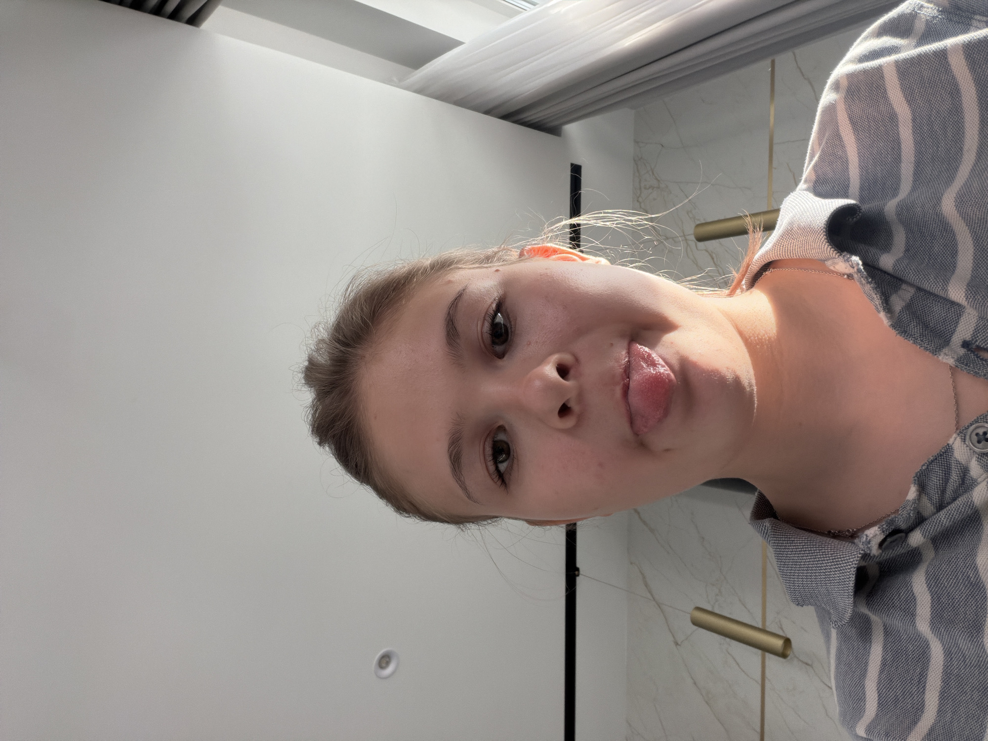 The image is a close-up, low-angle selfie of a young woman with light brown hair pulled back, sticking her tongue out. She is wearing a gray and white striped collared shirt and a silver necklace. Her face is illuminated by sunlight from the right. The background shows a modern interior with marble-patterned walls, a light fixture, and a window with sheer curtains. The ceiling is white with recessed lighting. There are no visible indicators of the location being Moscow, Russia.