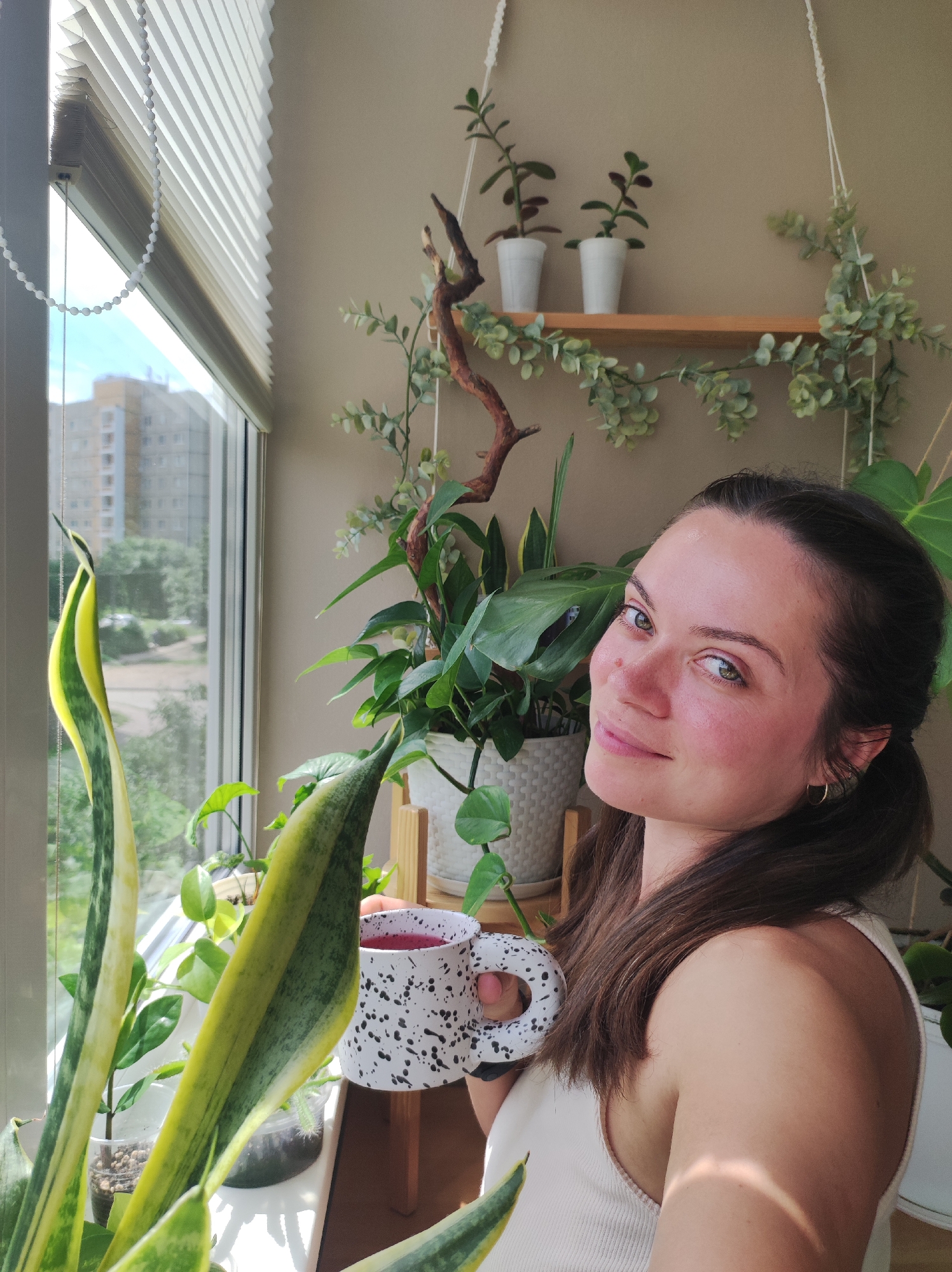 A young woman with brown hair is holding a mug of red liquid and looking at the camera. She is surrounded by several potted plants, some of which are hanging from the ceiling. The scene appears to be an indoor balcony or sunroom, with a large window that looks out onto a cityscape. Buildings and trees are visible through the window. The woman is wearing a light-colored tank top.