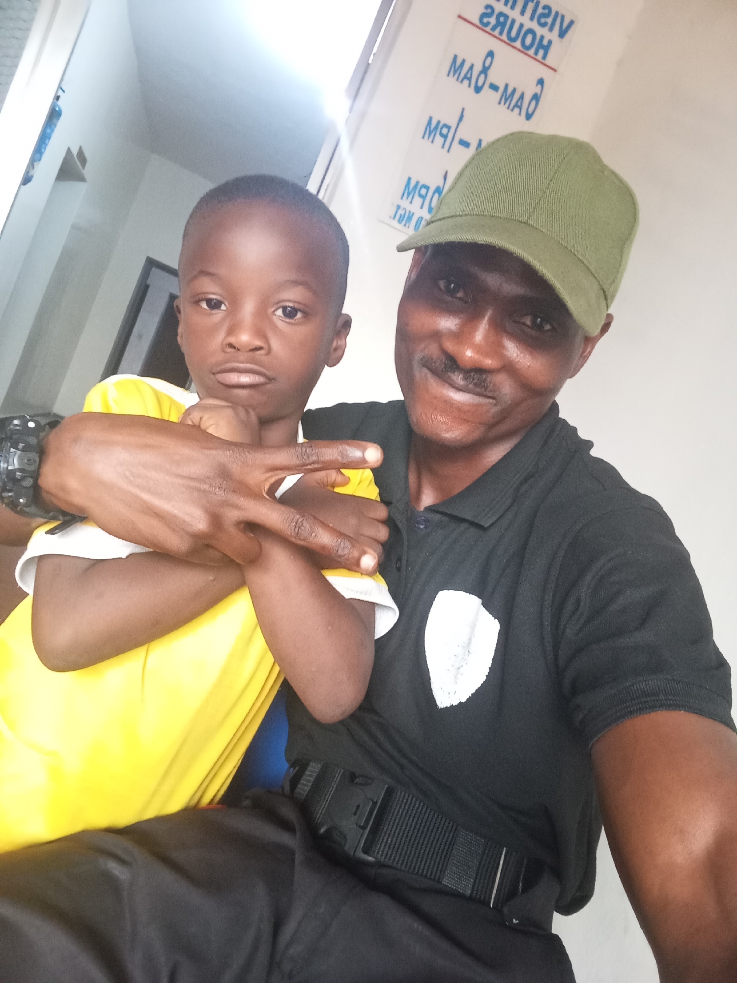 In this selfie, a man and a young boy are pictured together. The man, wearing a black polo shirt with a white emblem and a green baseball cap, has his arm around the boy. The boy, dressed in a bright yellow shirt, is making a peace sign with his hand, which is resting on his chest. They appear to be indoors, with a white wall and a sign in the background that reads "VISIT HOURS" in reversed lettering, suggesting it may be in a public or administrative building. The lighting suggests it could be daytime, possibly near a window. There are no specific visual cues that definitively place the location in Jos, Nigeria.
