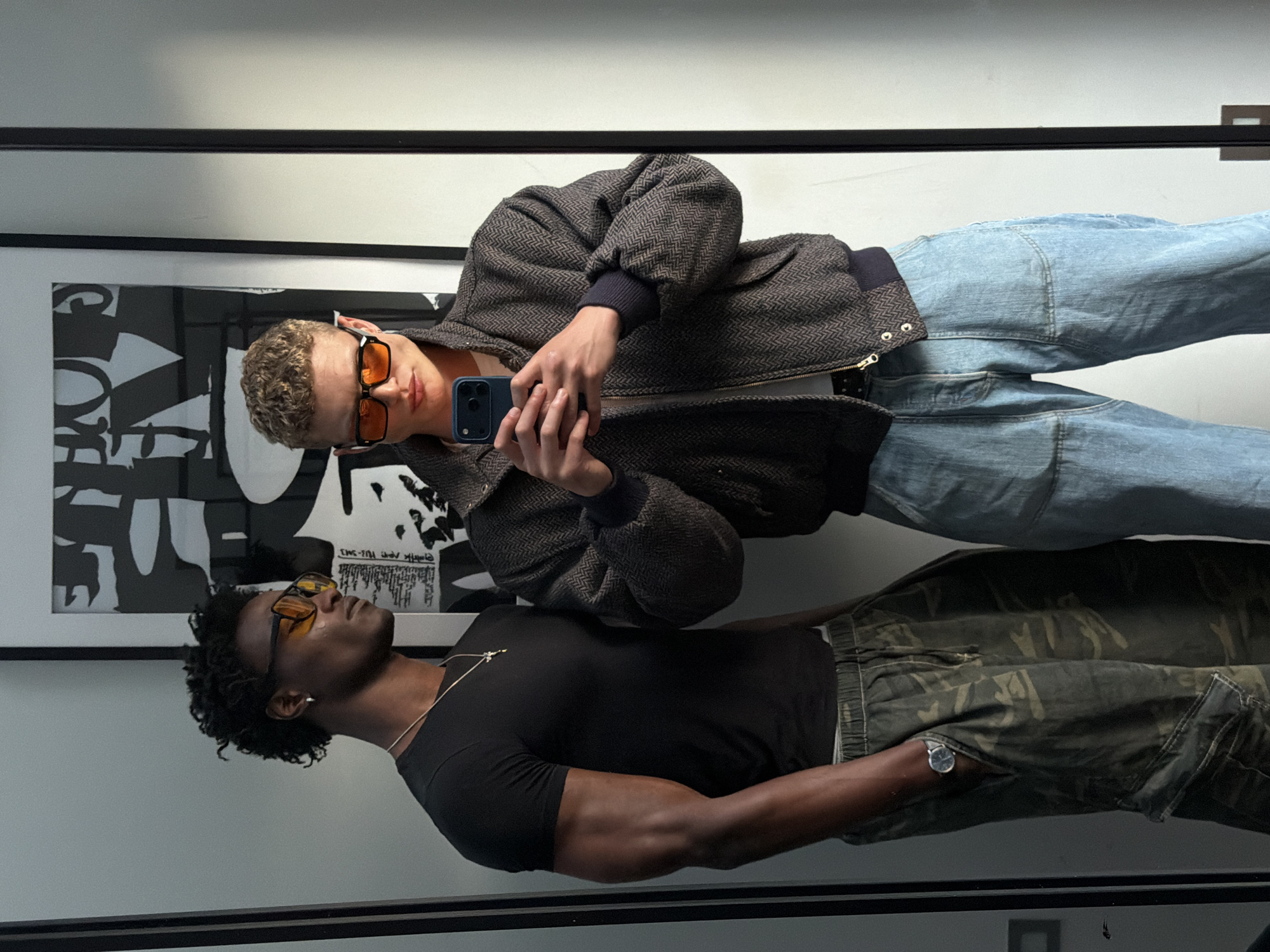 A medium shot depicts two individuals reflected in a mirror, positioned indoors against a wall. The upper reflection features a fair-skinned person with curly blonde hair and orange-tinted sunglasses, holding a smartphone and wearing a dark patterned jacket with light-wash denim jeans. The lower reflection shows a dark-skinned person with dark curly hair, wearing a black t-shirt, a silver chain necklace, a wristwatch, and camouflage pants. The background includes a black and white abstract print within a dark-framed picture hanging on the wall. The setting appears to be an interior space, likely a retail environment or residence. Location in London, United Kingdom, cannot be determined.
