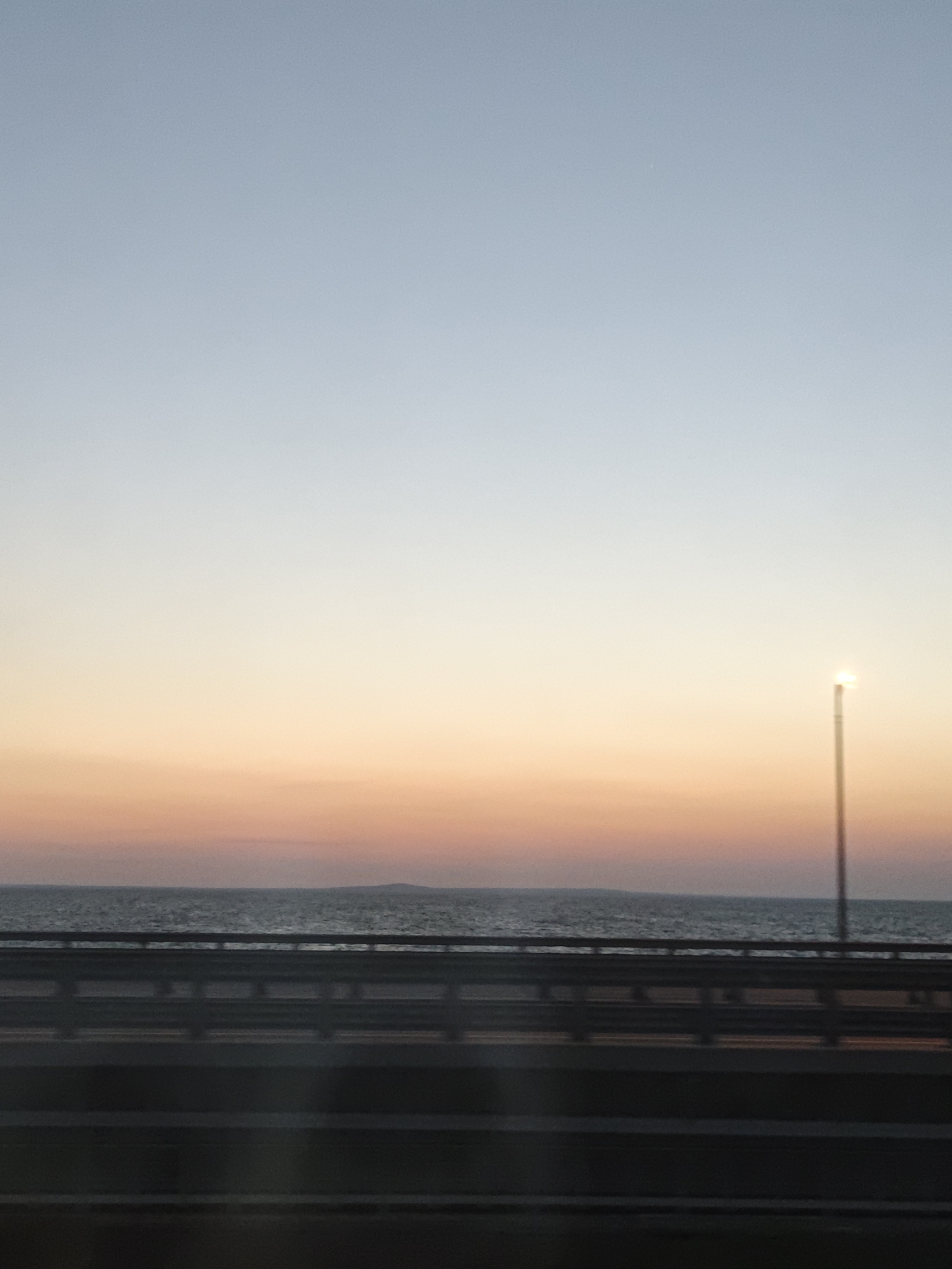 The image shows a serene sunset over the sea, viewed from a bridge or elevated roadway. The sky transitions from a pale blue at the top to soft hues of orange and pink near the horizon. A solitary lamppost stands on the right side, its light reflecting a bright glow in the dusky sky. Below the sky is a calm sea, with a faint, dark line of land or a distant island visible on the horizon. The foreground is dominated by the structure of the bridge, with dark railings and horizontal lines suggesting multiple levels or lanes. The overall impression is one of tranquility and the end of a day. The image is taken in Yalta, Ukraine, likely from the Yalta Viaduct or a similar elevated structure overlooking the Black Sea.