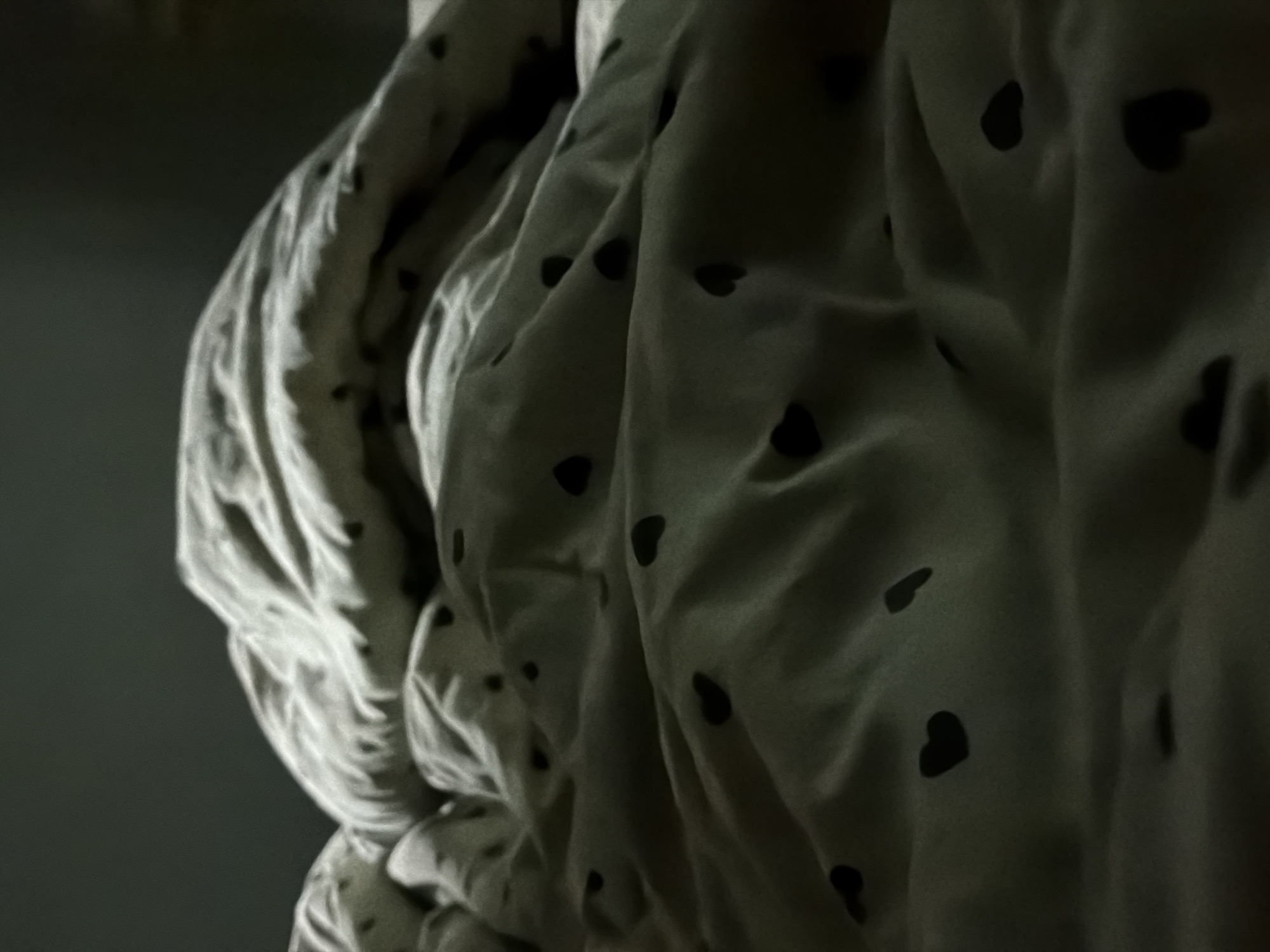 The image shows a rumpled white bed sheet with small black heart patterns. The sheet is draped in multiple folds, creating highlights and shadows on the fabric. The background appears to be a plain, dark wall, suggesting the photo was taken indoors. No people are visible. The location in the city of Chaikovsky, Russia is not apparent from the visual information.