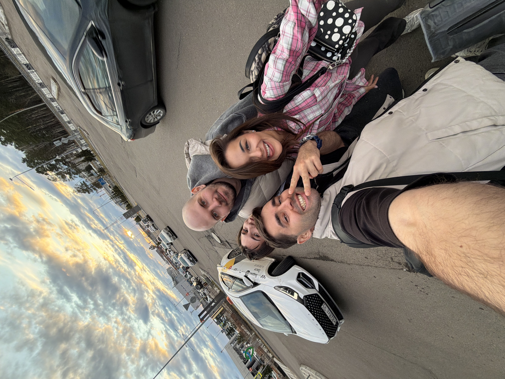 A group of four individuals is captured in a selfie-style photograph taken in Емельяново Airport, Russia. The photo is oriented at an angle, showing a section of the airport parking lot with cars and a sky filled with clouds at sunset. The people are posing close together. One of them is a bald man with a grey hoodie. Beside him is a woman with brown hair, wearing a pink plaid shirt and black pants. Another man to the right is making a peace sign with his fingers, sporting a white jacket. The person taking the photo is presumably a man with visible arm hair in the foreground. The background shows cars lined up in the parking area, including a white car with "taxi" markings. The sky transitions from a bright sunset to a darker evening hue, with golden-colored clouds.