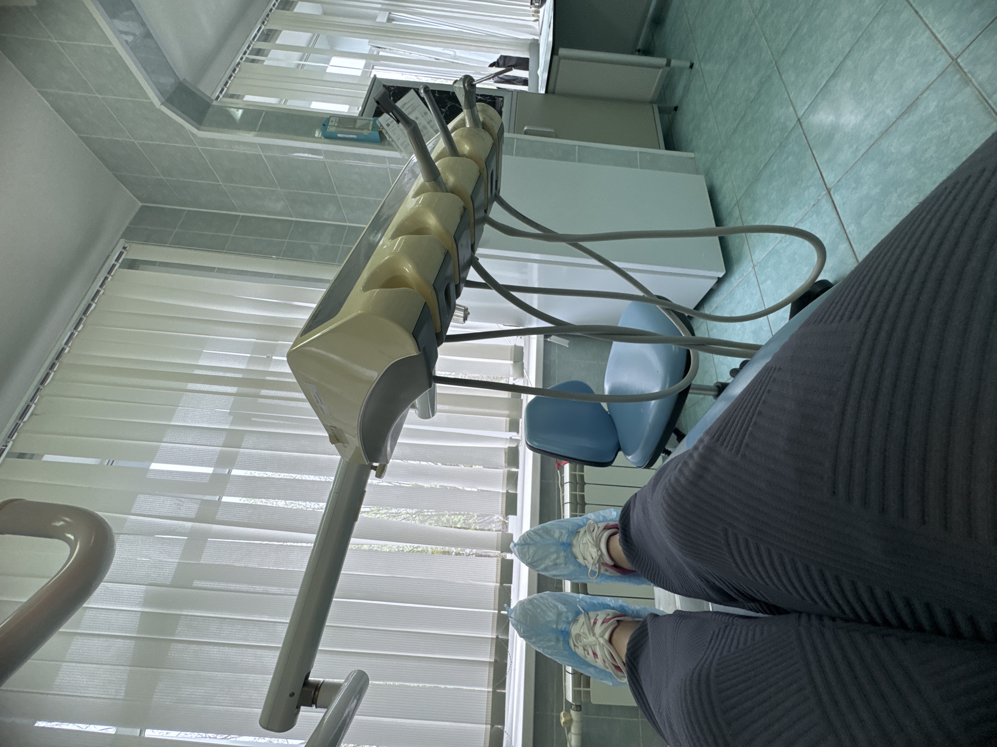 A patient in a dental chair wearing protective shoe covers in a dental office in Zubovo, Russia. The dental unit is visible on the left, with various dental tools and hoses. A blue dental chair is positioned next to the unit. Window blinds are in the background. The walls are tiled. The patient is wearing dark trousers.