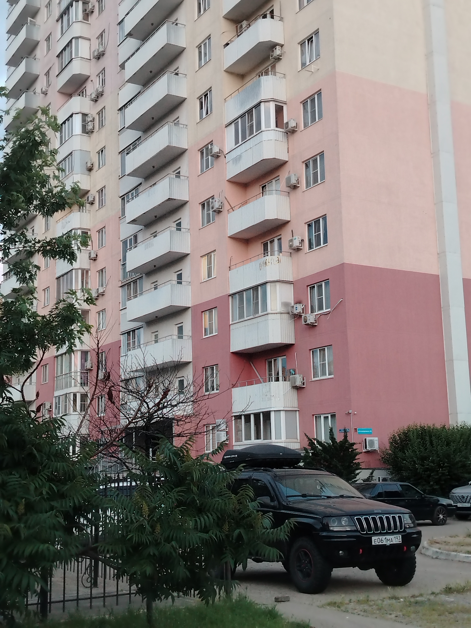 A black Jeep Grand Cherokee with a roof cargo box is parked on a paved surface in front of a multi-story apartment building. The building has a light beige and pink color scheme, with numerous balconies. Trees and greenery are visible in the foreground and to the right of the vehicle. The license plate of the Jeep reads "E061MA 193". A black sedan is parked behind the Jeep, and a silver car is partially visible to the right of the sedan. In the background, street signs are visible on the building. This scene is located in Krasnodar, Russia.