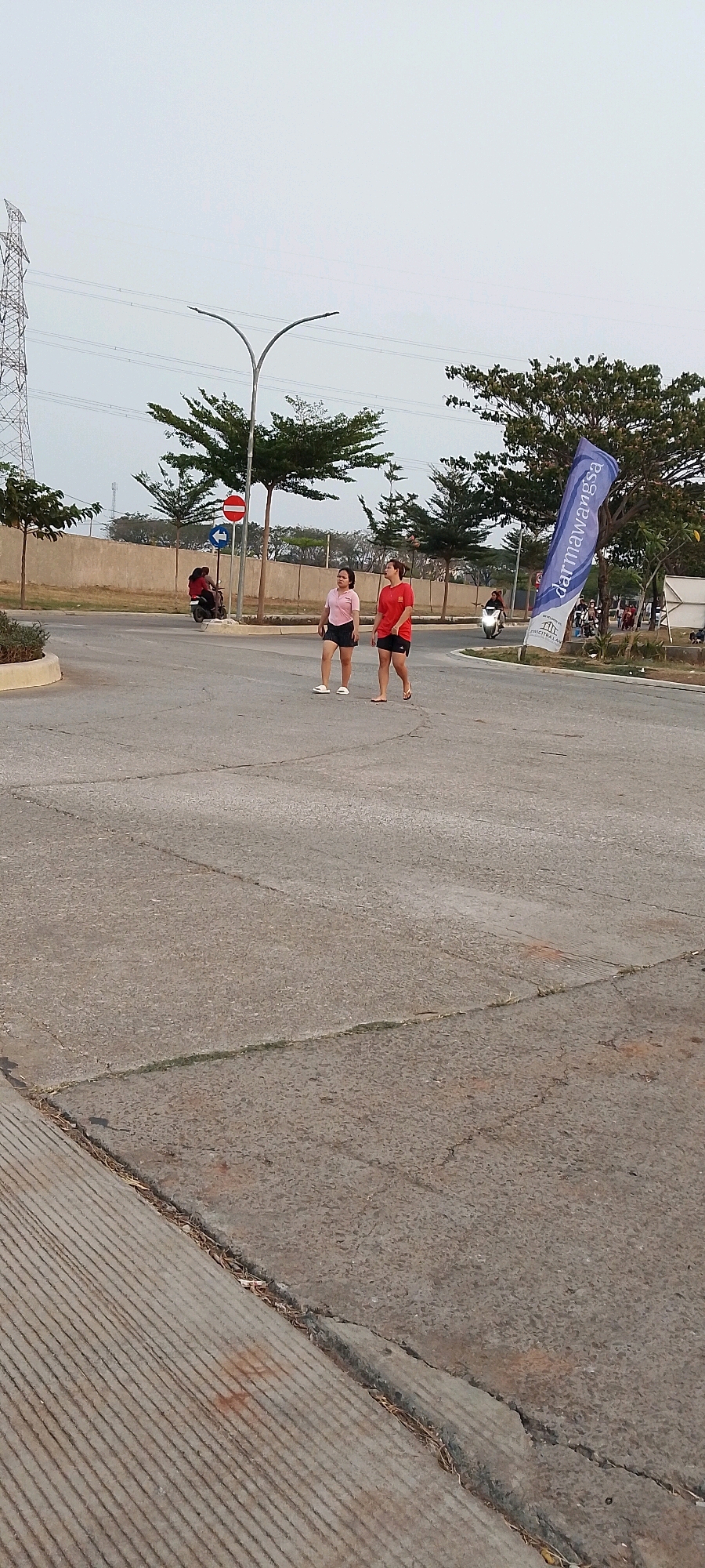 Two young women, one wearing a pink t-shirt and black shorts, and the other in a red t-shirt and black shorts, are walking side-by-side on a paved road. The woman in pink is to the left, and the woman in red is to the right. They are in an outdoor environment, possibly in the city of Satriamekar, Indonesia, as indicated by the presence of a tall electrical pylon and various trees. The road appears to be asphalt, with some cracks and debris. In the background, a person is riding a motorcycle. A blue and white flag with text is visible on the right side of the image. The sky is overcast and light.