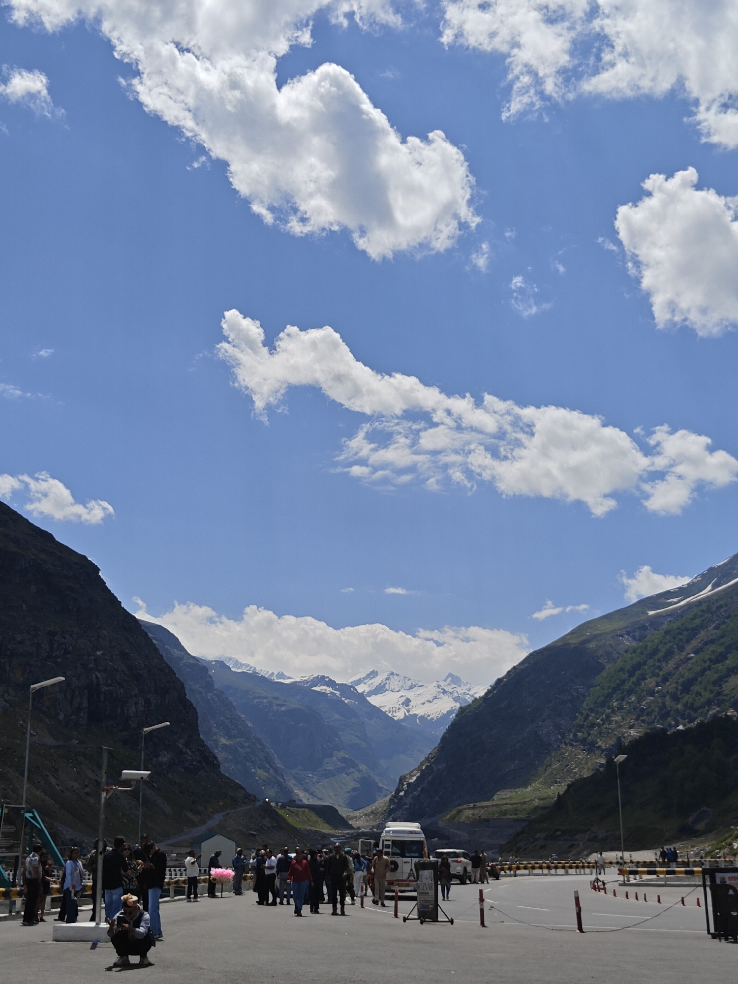 A vertical, eye-level shot captures a vibrant scene in a mountainous region of India, possibly near Rohtang Pass, with a backdrop of snow-capped peaks under a bright blue sky dotted with fluffy white clouds. The foreground features a paved area with several people gathered, some standing, some walking, and one person crouched down, looking at their phone. Various vehicles, including a white van, are parked or driving on a road that curves into the distance. The sides of the valley are lined with steep, dark, rocky mountains on the left and green, vegetated slopes on the right. Streetlights are visible along the road.