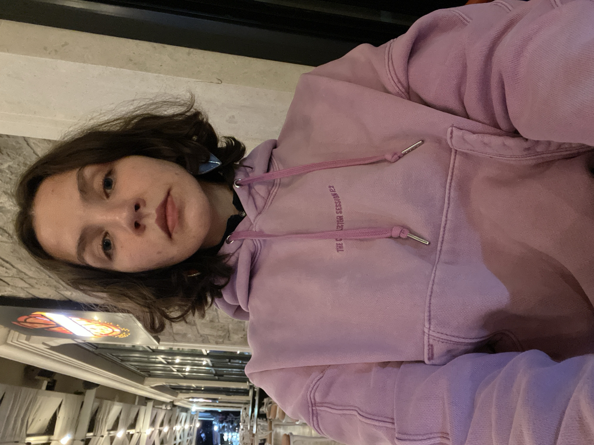A young woman with shoulder-length brown hair is posing for a selfie in an outdoor setting. She is wearing a lavender-colored hooded sweatshirt with drawstrings. She is slightly angled, with her face tilted towards the camera. The background shows an outdoor dining area with string lights hanging above tables. A sign with a pizza graphic is also visible. Based on the location, it appears to be an outdoor restaurant or cafe in Bečići, Montenegro.