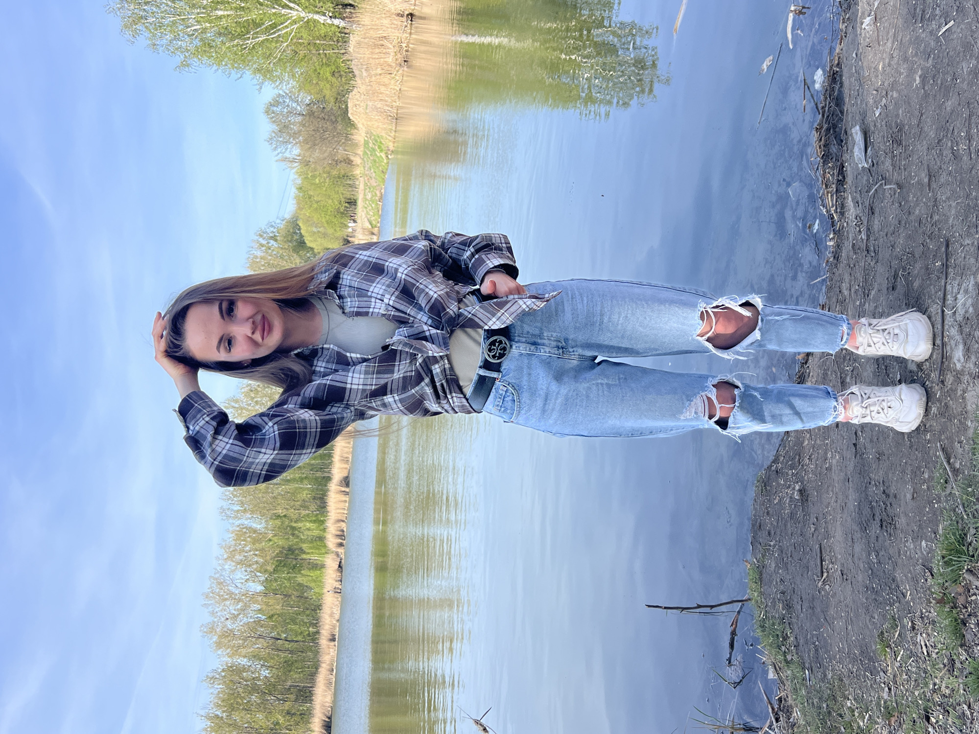 A young woman stands on the edge of a body of water, possibly a lake or river, in Ciemne, Poland. She is wearing ripped light blue jeans, a grey/black flannel shirt, white sneakers, and a dark belt with a large buckle. She is posing, with one hand on her head and the other in her pocket. The water reflects the trees and sky. The immediate ground is earthen, with some trash and vegetation.