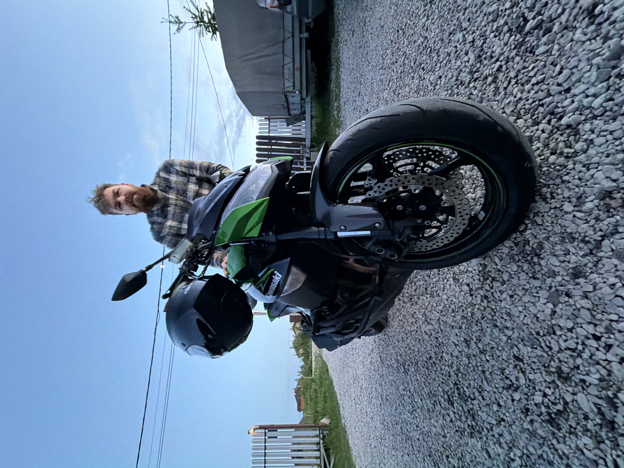 The image shows a man and a black and green Kawasaki motorcycle on a gravel driveway in Khutyn', Russia. The man, with a beard and wearing a plaid shirt, is partially visible behind the motorcycle. A black helmet hangs from the motorcycle's handlebars. The background includes a gray trailer, a wooden fence, and electrical lines against a blue sky. The ground is covered in light gray gravel.